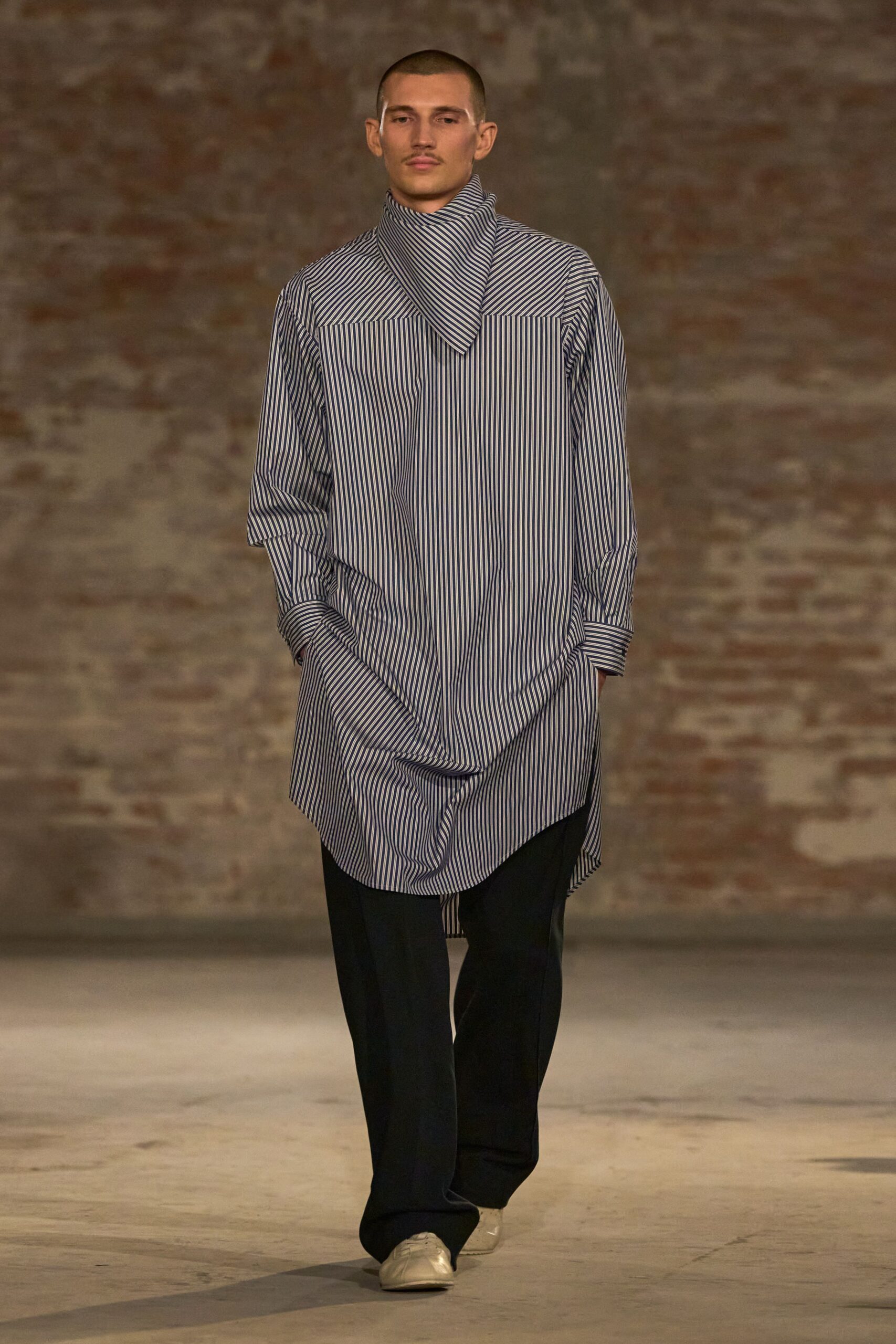 Blue-and-white banker stripe cotton, with a scarf panel crossing into a high funnel neck. Black straight trousers, breaking softly over the shoes. Cream leather lace-ups with blunt rounded toes. Crossing a museum courtyard after rain, map folded in one pocket.