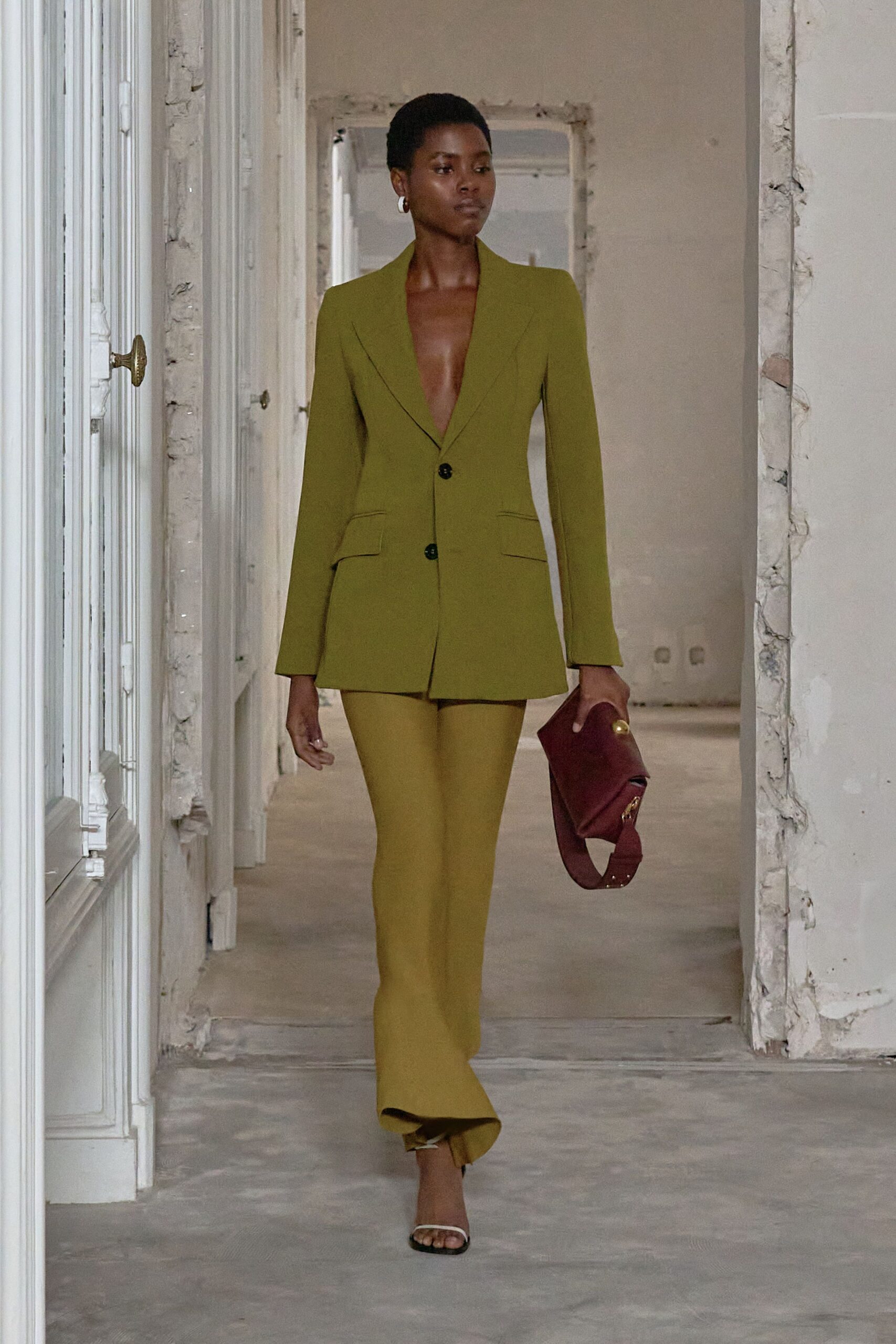 Olive tailored blazer worn bare, plunging shawl lapels and two dark buttons. Chartreuse slim flares breaking softly over the front of the sandal. Burgundy folded leather clutch with a wide strap and gold stud. White barely-there sandals. Walking through an unfurnished apartment before the keys change hands.