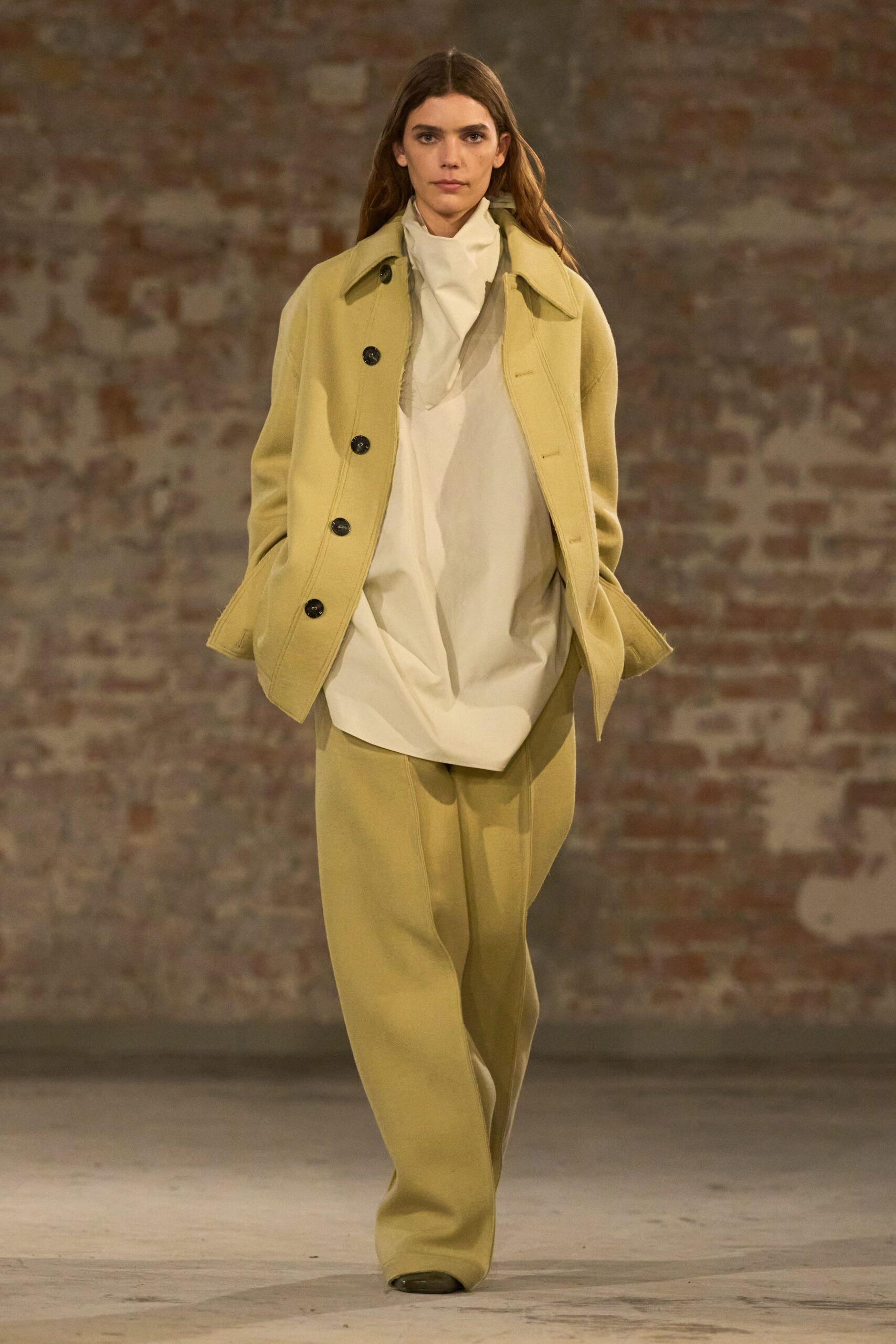 Pistachio wool coat with oversized black buttons and dropped shoulders. Ivory high-neck blouse cut long and bloused into a soft front fold. Matching wide-leg trousers with a pressed crease breaking over the shoe. Early train to the coast, thermos still too hot to drink.