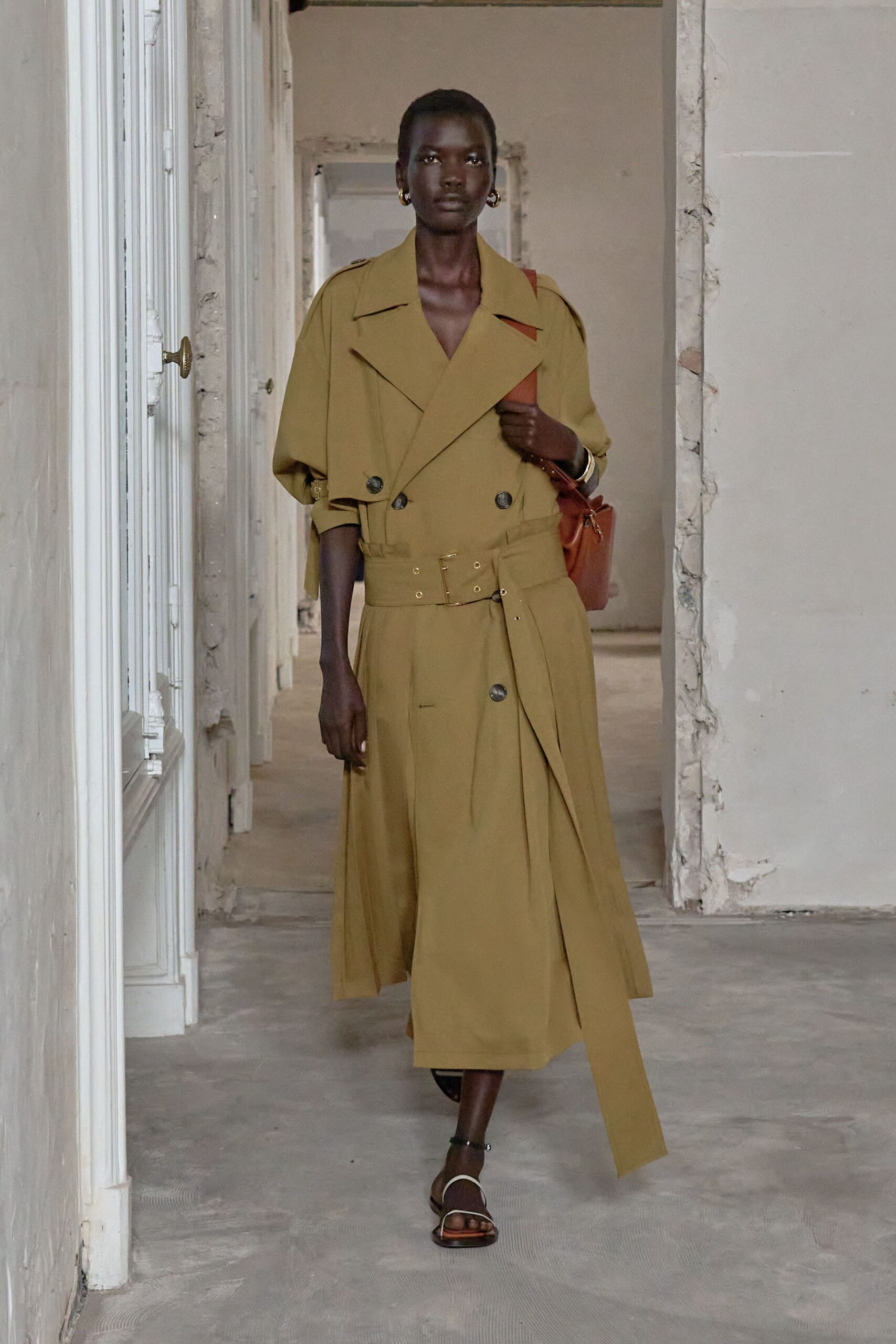 Ochre double-breasted trench dress with dropped sleeves and a wide grommet belt. Cognac leather shoulder bag tucked close under the arm. Small gold hoops. Stacked gold bangles. Black ankle-strap sandals with a pale curved toe band. Walking through a half-renovated apartment before the movers arrive.