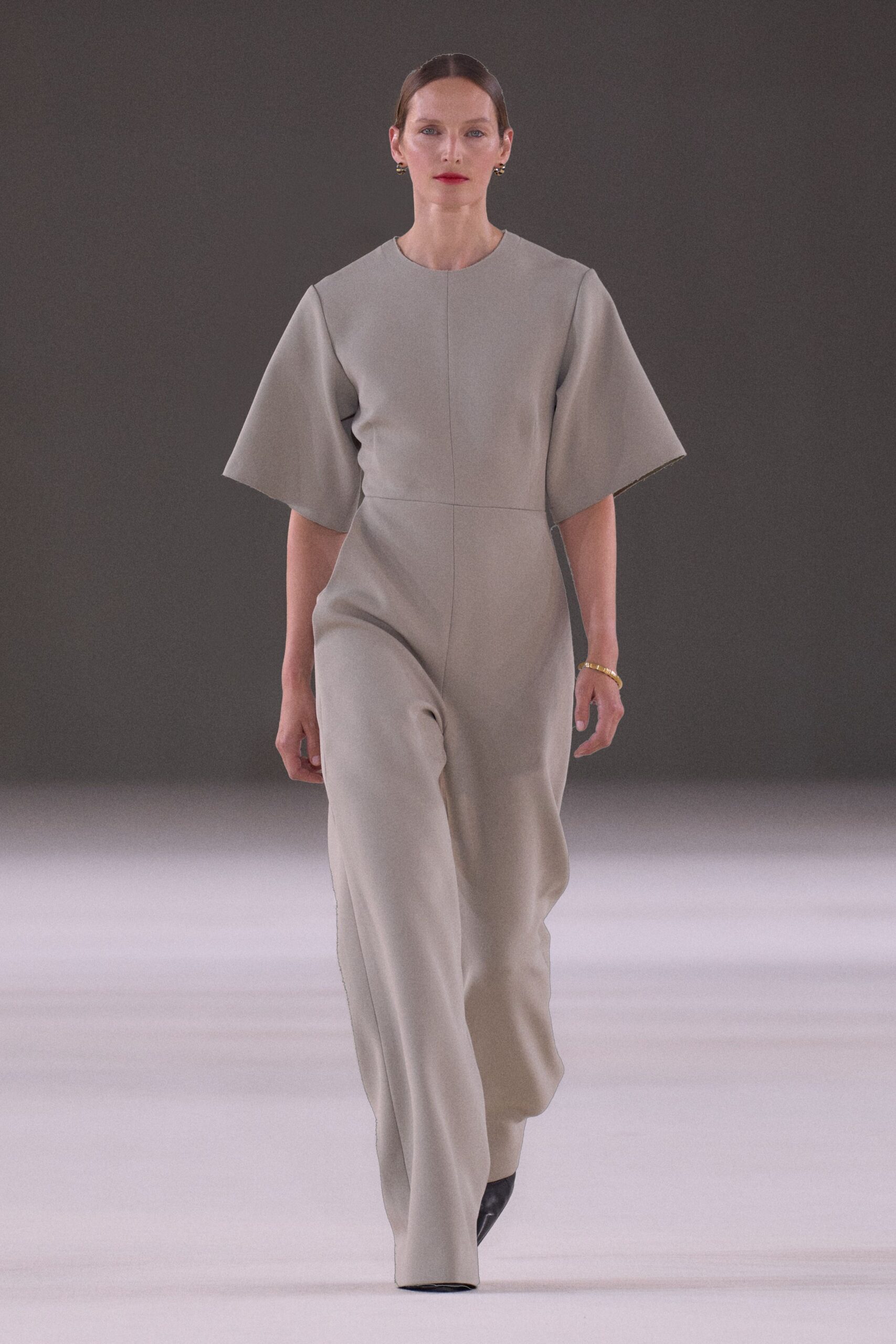 Pale dove jumpsuit in dense crepe, cut with wide bell sleeves and a clean vertical seam. Small gold knot studs. Ridged gold bangle at one wrist. Black pointed boots just breaking the hem. For crossing a museum atrium before the doors open to the public.