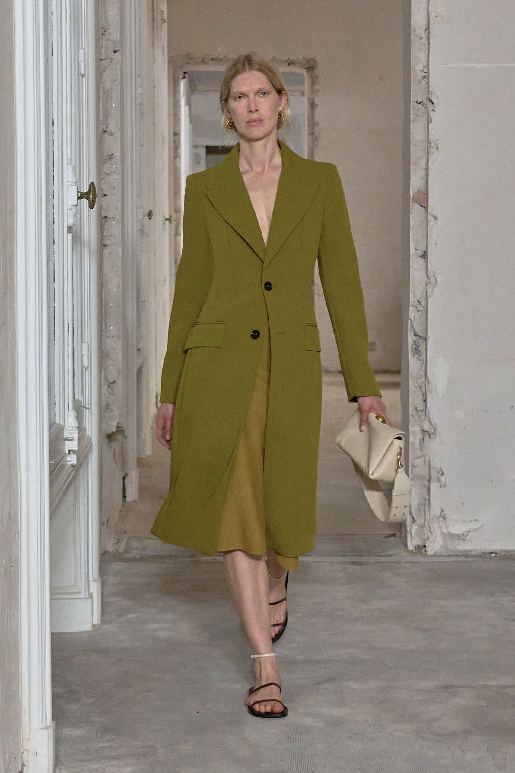 Olive tailored coatdress with a plunging lapel and two black buttons. Moss satin slip skirt peeking from the parted hem. Small twisted gold hoops. Soft ivory clutch, folded under a wide studded strap. Barely there black ankle-strap sandals. Walking through an unfurnished apartment before the plaster dust settles.