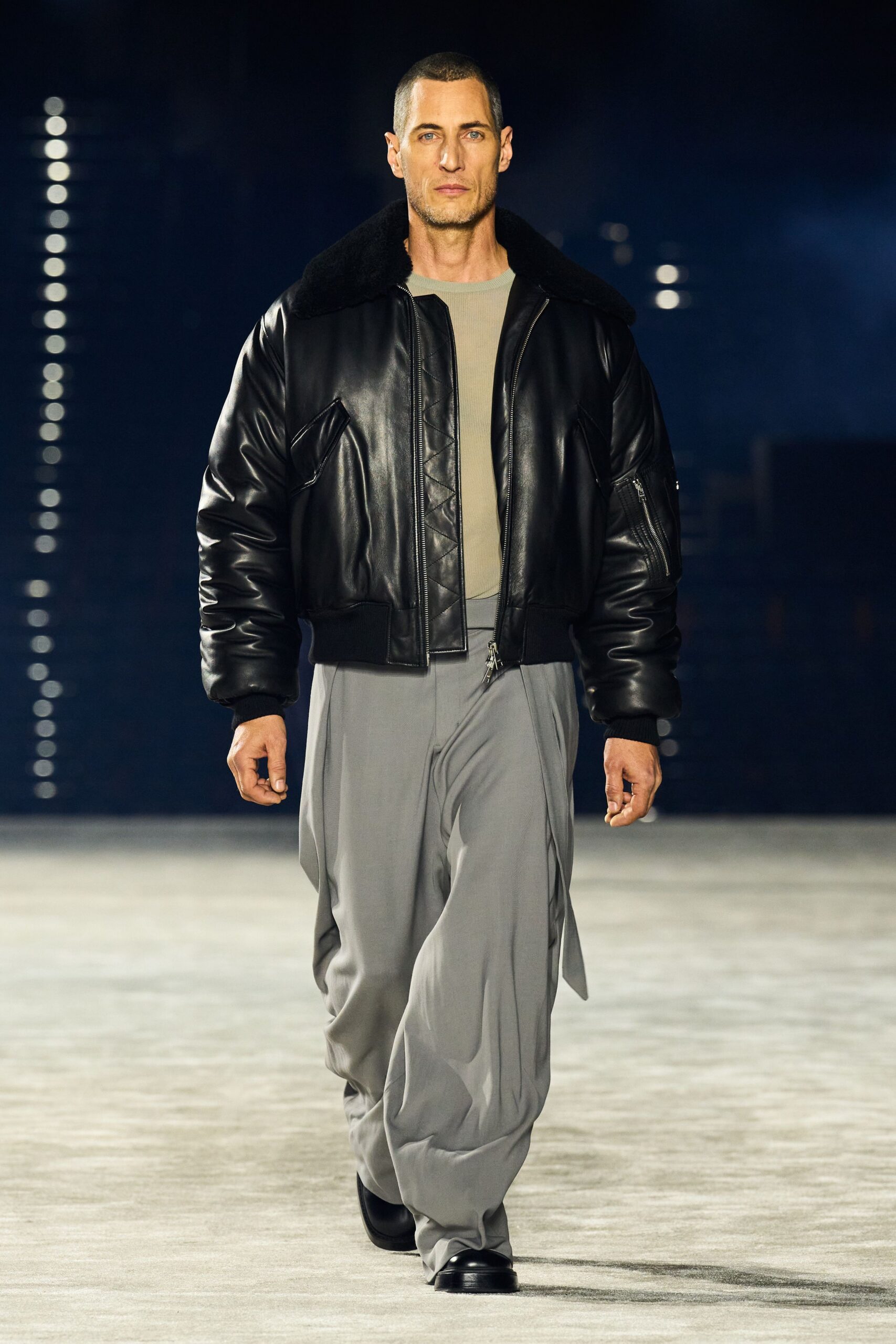 Black leather bomber with a deep shearling collar and sleeve zip pocket. Pale sage ribbed tee cut close at the neck. Pearl-grey fluid trousers, long hems collapsing over the shoes. Black leather square-toe shoes. Walking the hotel corridor at dawn before the conference coffee is set out.
