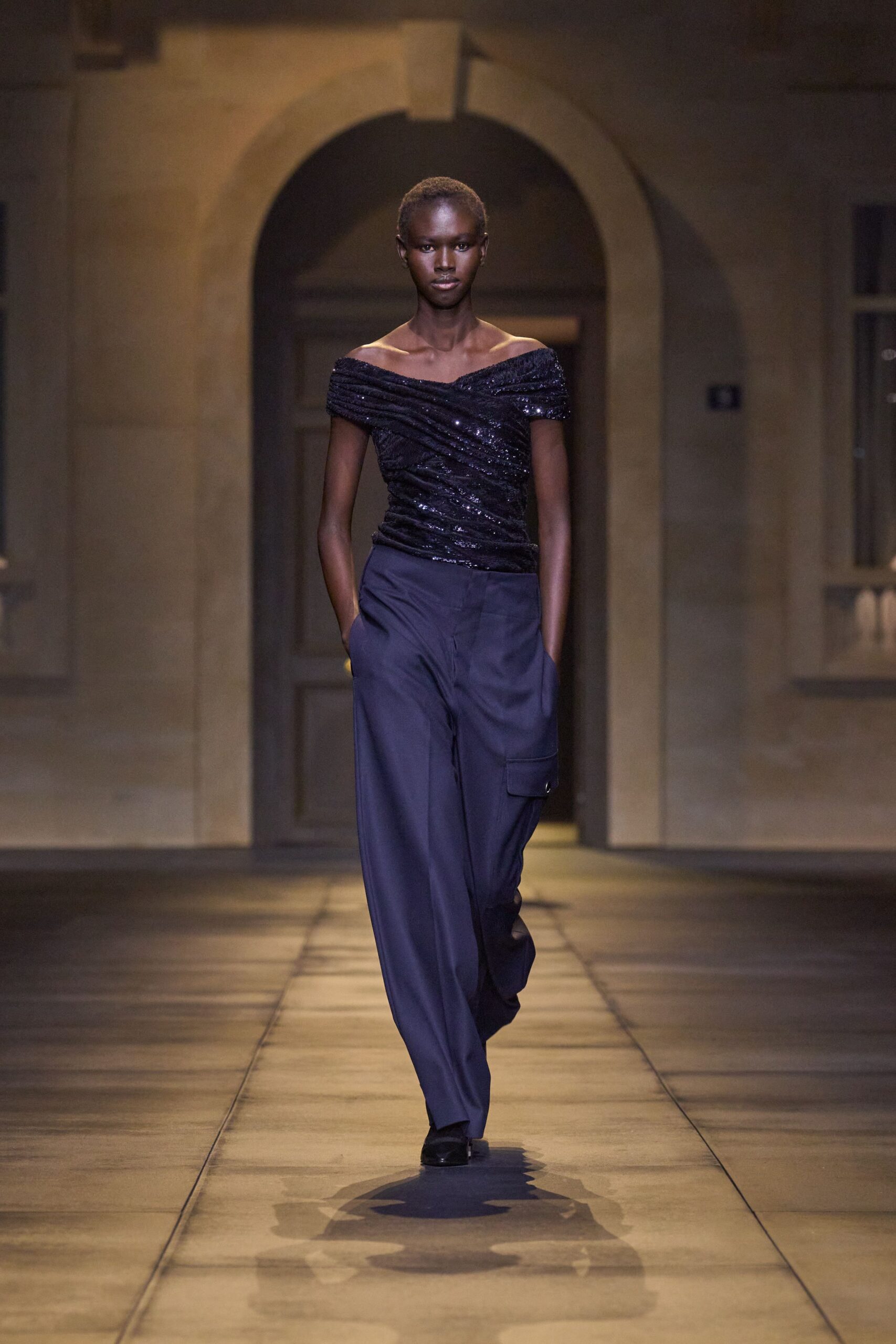 Ink-black sequined top, ruched diagonally into an off-shoulder band. Midnight wide-leg trousers with a lone flap pocket on the thigh. Black square-toe shoes, barely breaking the hem. Crossing a stone courtyard after midnight, music still trapped in the walls.