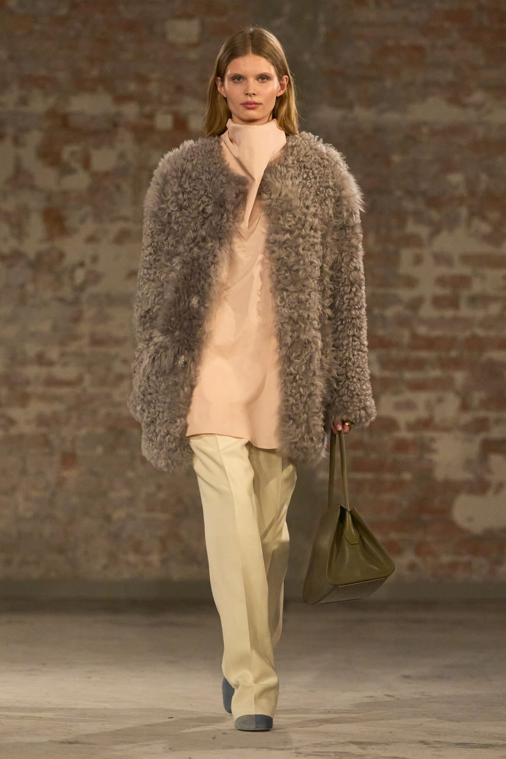 Taupe curly shearling coat, cut collarless and worn hanging open. Blush crepe tunic with a tall funnel neck and deep slit. Cream tailored trousers with a pressed crease breaking over the shoe. Olive leather trapeze tote on slim shoulder straps. Grey suede pointed boots. Early hotel checkout, lobby flowers already being changed.