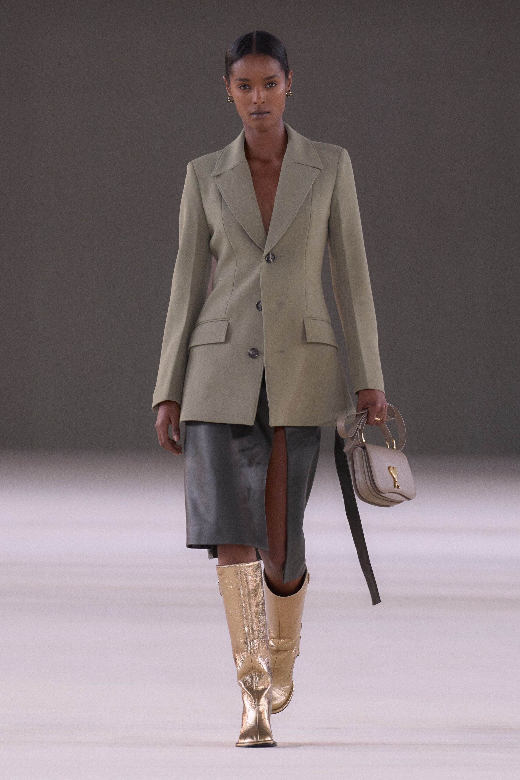 Sage tailored blazer cut long, with wide lapels and dark horn buttons. Black leather midi skirt, split high at the front. Taupe saddle bag with a clear looped top handle. Crinkled gold knee boots with square toes. For crossing a hotel lobby before the bar opens at noon.