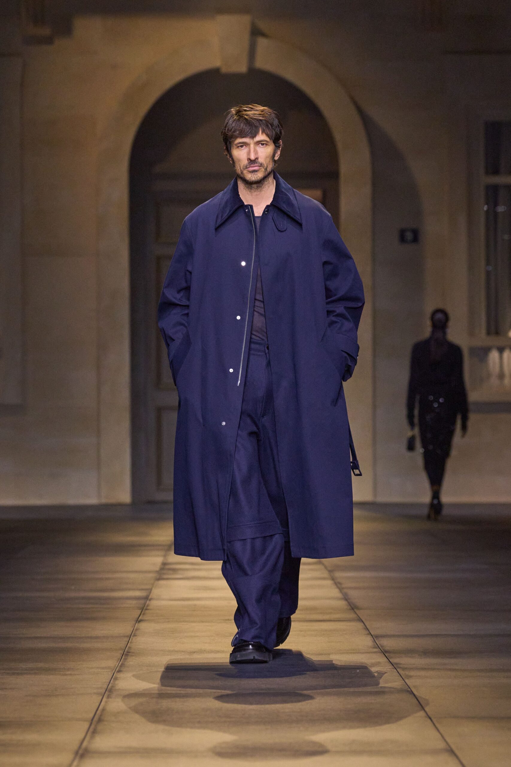 Inky navy overcoat with a long silver zip and wide pointed collar. Sheer midnight crewneck peeking through the opening. Fluid navy trousers breaking softly over the shoe. Black leather slip-ons with a blunt square toe. Crossing a stone courtyard just before the museum opens.