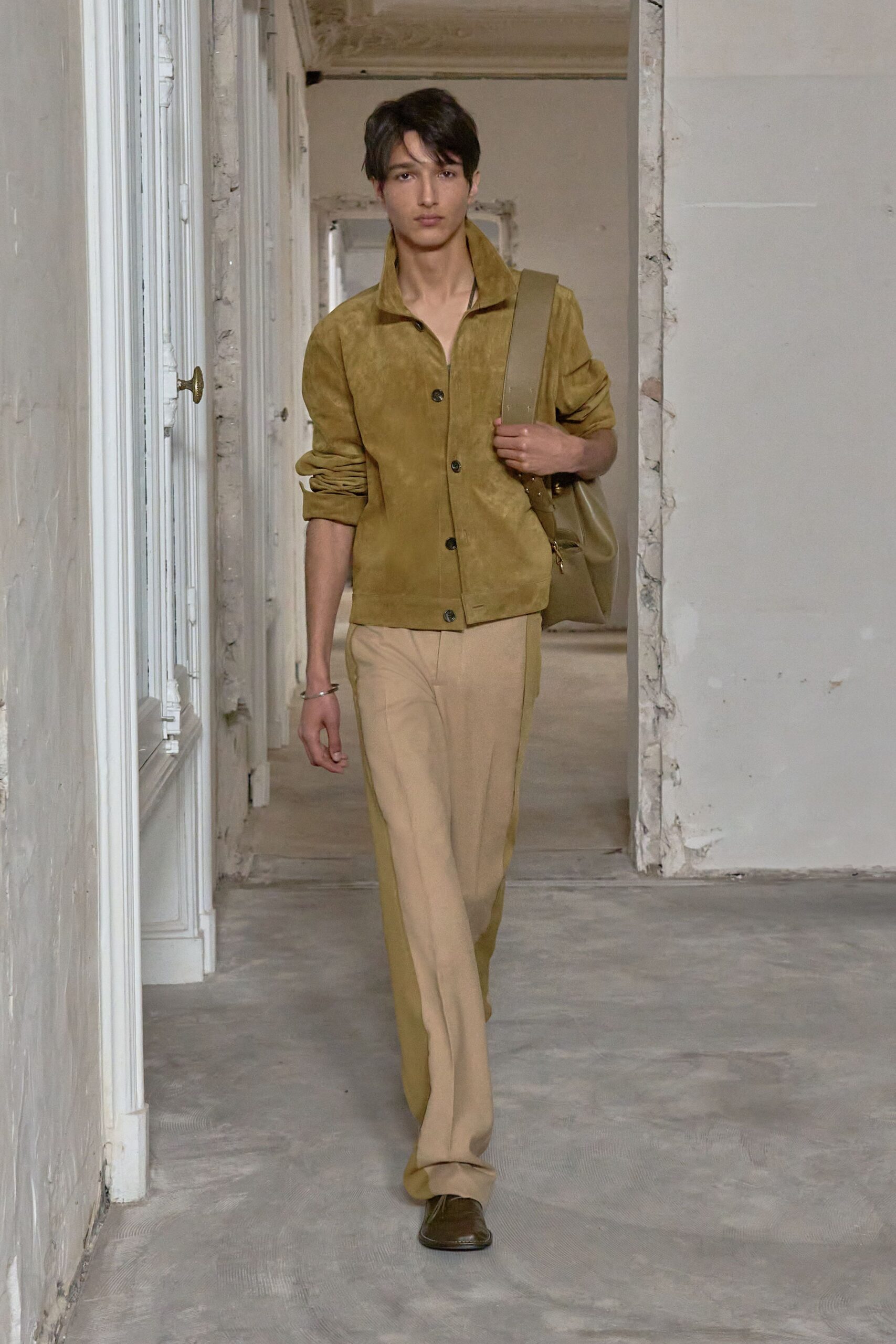 Moss suede jacket with a high stand collar and dark horn buttons. Taupe leather hobo bag on a wide strap. Sand trousers with a pressed crease and extra length pooling lightly. Olive leather square-toe shoes. Thin silver bangle. Walking through an unfurnished apartment before the keys change hands.