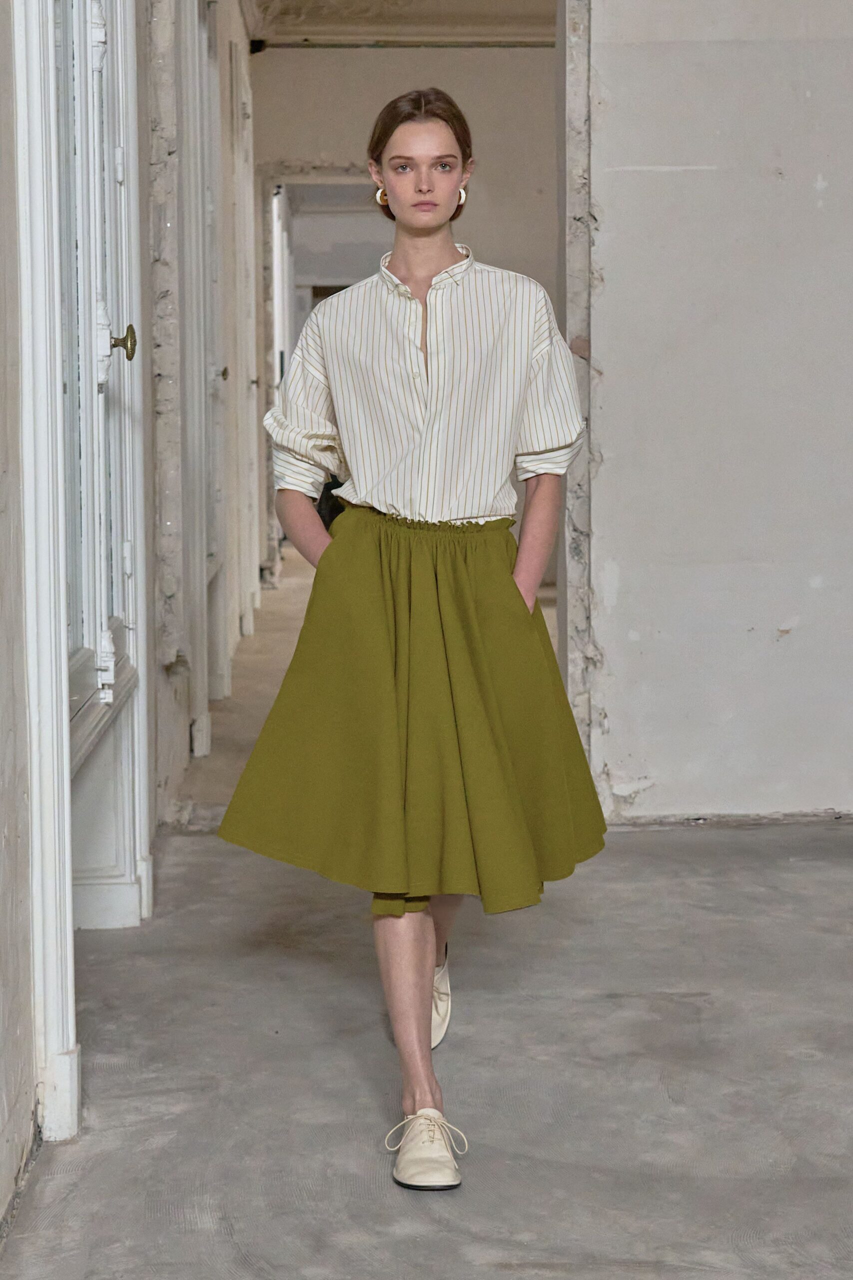 Mismatched ivory hoops hugging the lobes. Cream cotton shirt with ochre banker stripes and a soft band collar. Olive gathered midi skirt, full and belling from a ruched waist. Cream lace-up flats with blunt toes. Sunday museum shift, keys jangling somewhere in the pockets.
