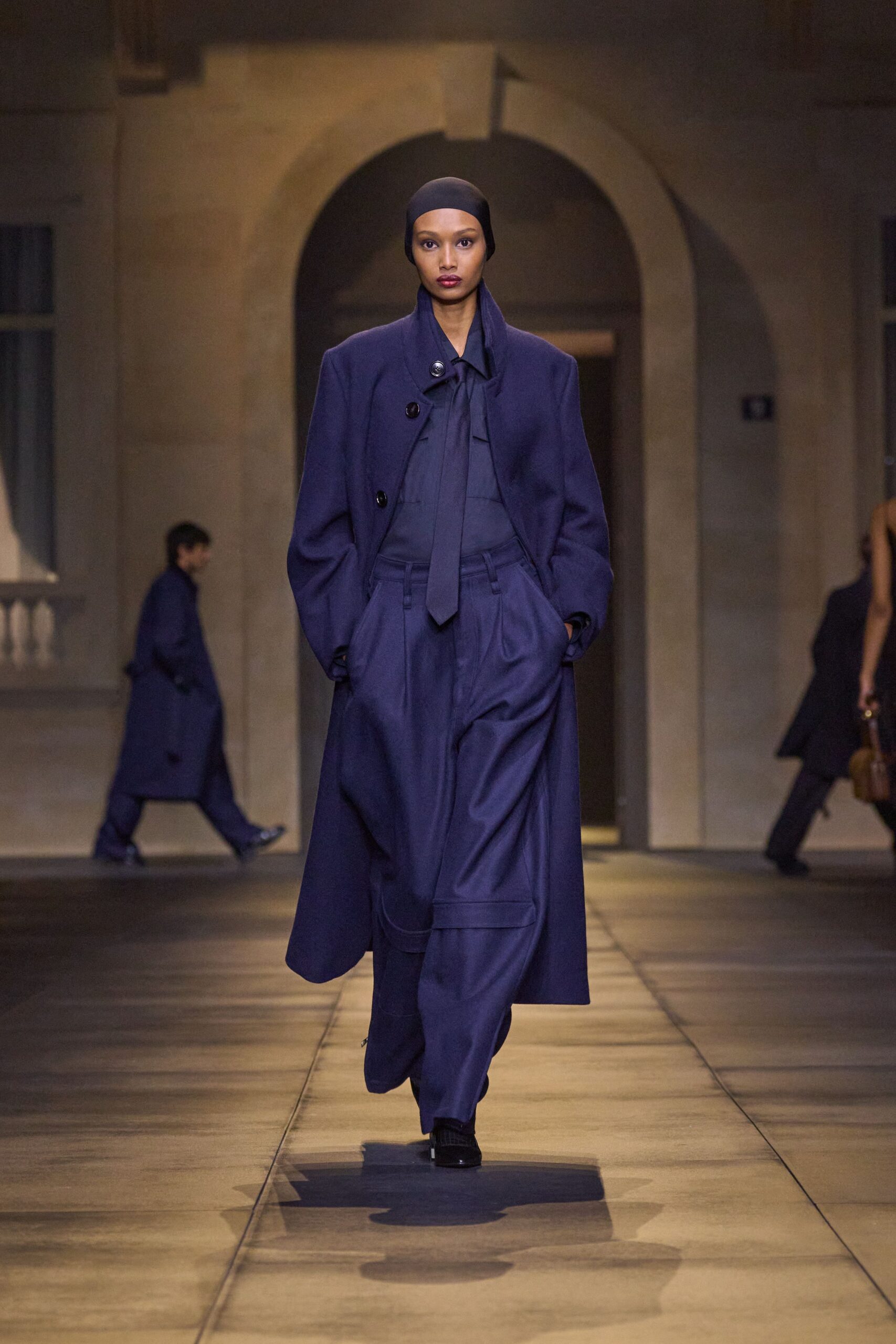 Black stretch cap pulled smooth over the ears. Inky wool coat with squared shoulders and off-center glossy buttons. Tone-on-tone navy shirt with a long tie hanging straight. High-waisted pleated trousers, cut full through the leg. Black square-toe shoes. Crossing a stone courtyard just after the rain stopped.