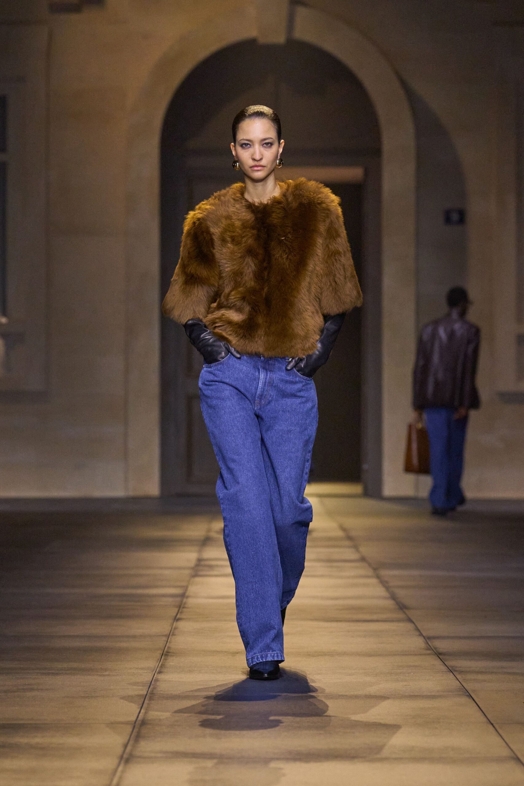 Burnt-caramel fur top with a rounded hem and broad elbow sleeves. Black leather gloves disappearing into the pockets. Mid-blue straight jeans, pressed crease held down the front. Dark gold orb earrings. Black pointed shoes. Crossing a stone courtyard just before the museum doors unlock.