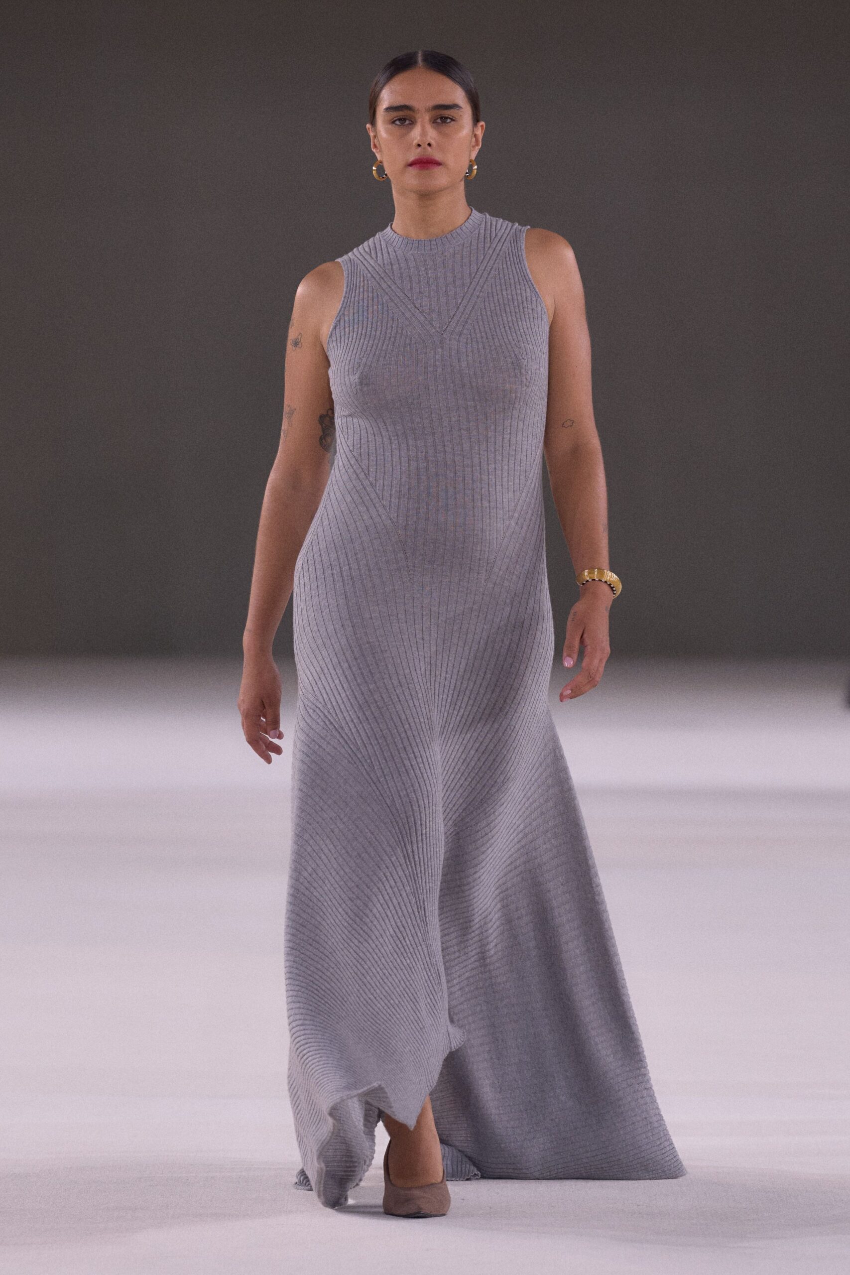 Pale grey rib-knit maxi, engineered seams fanning from the mock neck. Small gold hoops. Wide gold link bracelet. Taupe pointed flats under the swaying hem. Crossing a museum atrium before the air-conditioning fully wakes up.
