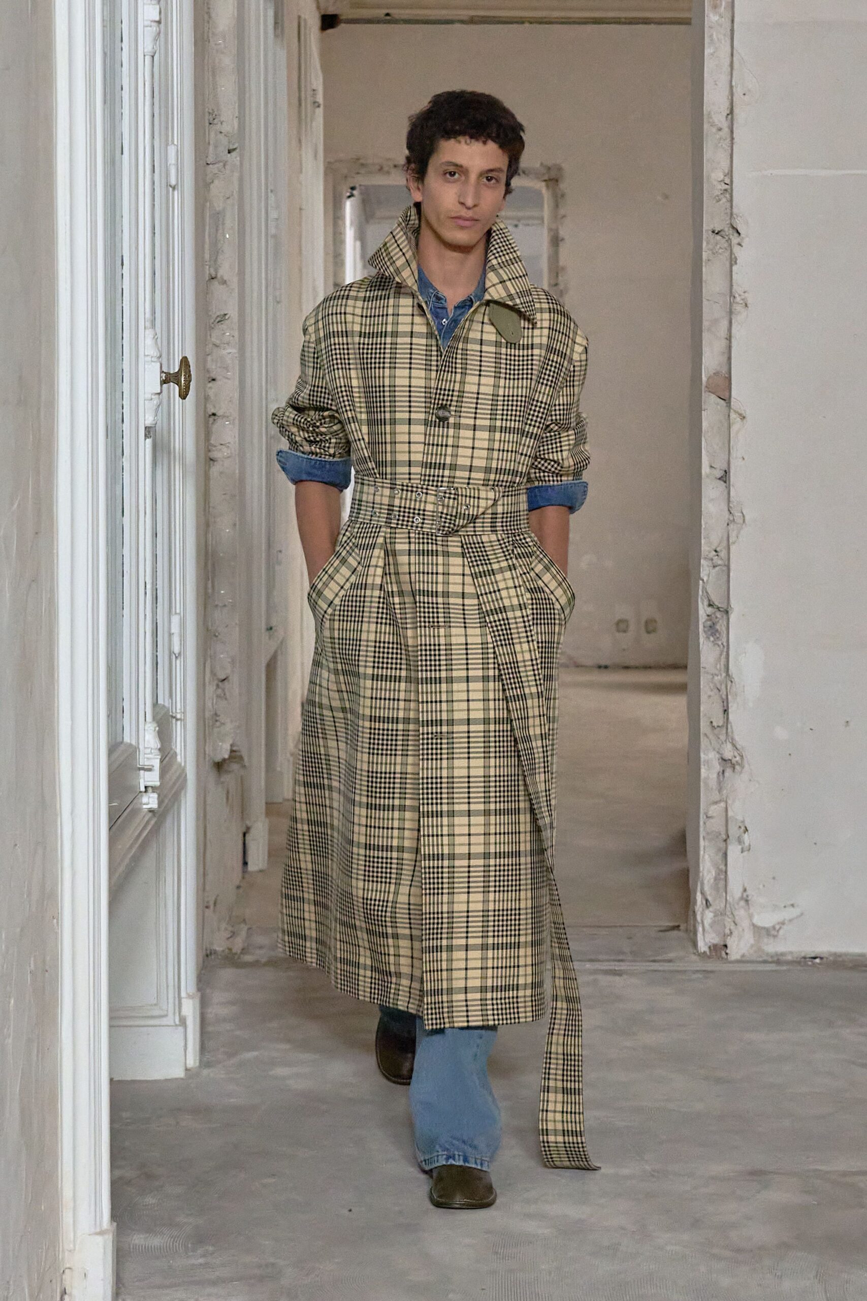 Cream tartan trench cinched with a grommeted self-belt hanging past the hem. Faded blue chambray shirt, sleeves rolled thick above the elbow. Pale blue jeans, straight through the leg. Dark square-toe boots. Walking through a half-renovated apartment before the radiator heat comes on.