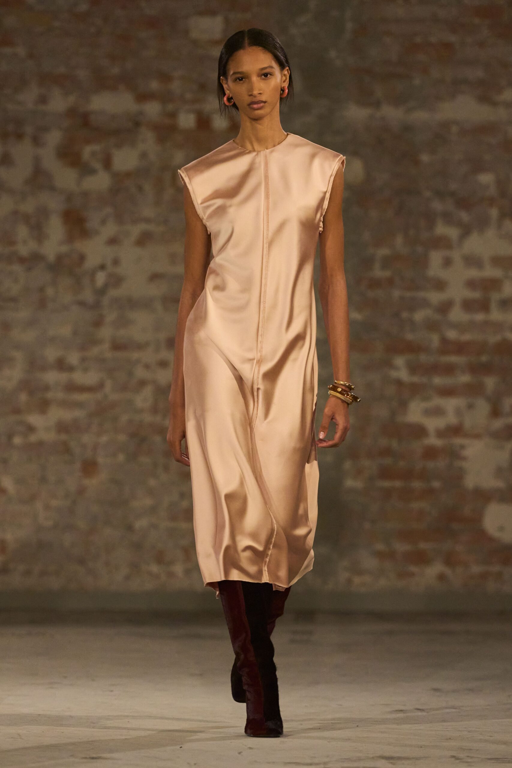 Blush satin column dress with raw cap sleeves and a raised center seam. Coral hoops at both ears. Stacked mixed-metal bangles on one wrist. Oxblood boots, velvety nap catching the light. Early dinner in a half-renovated palazzo, plaster dust still in the air.