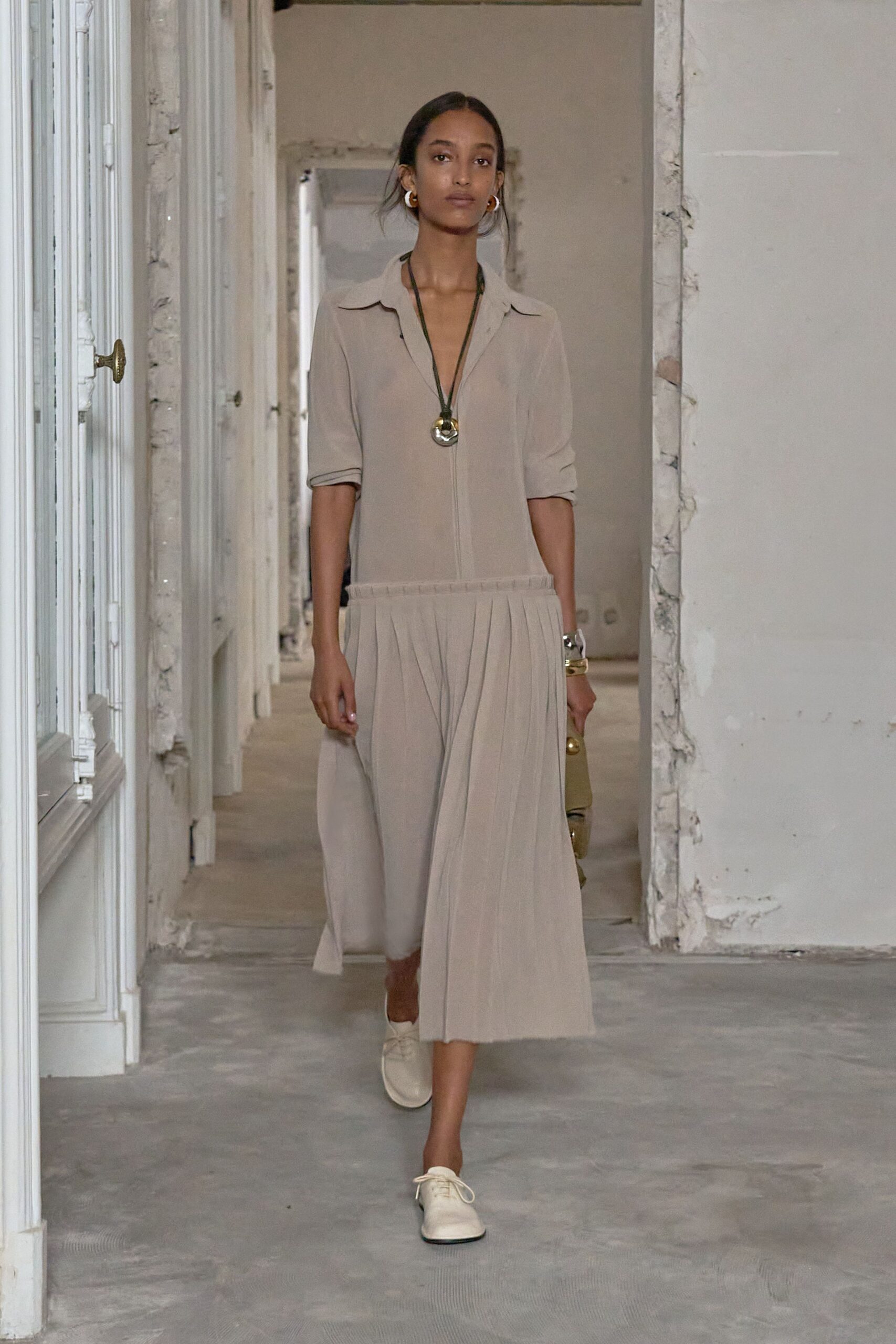 Pale greige shirt dress in fluid crepe, dropped waist falling into knife pleats. Oversized gold disc pendant on a dark cord. Thick gold hoops. Wide gold cuff stacked with a clear bangle. Cream lace-up flats with squared toes. Walking through a half-renovated apartment before the first furniture arrives.