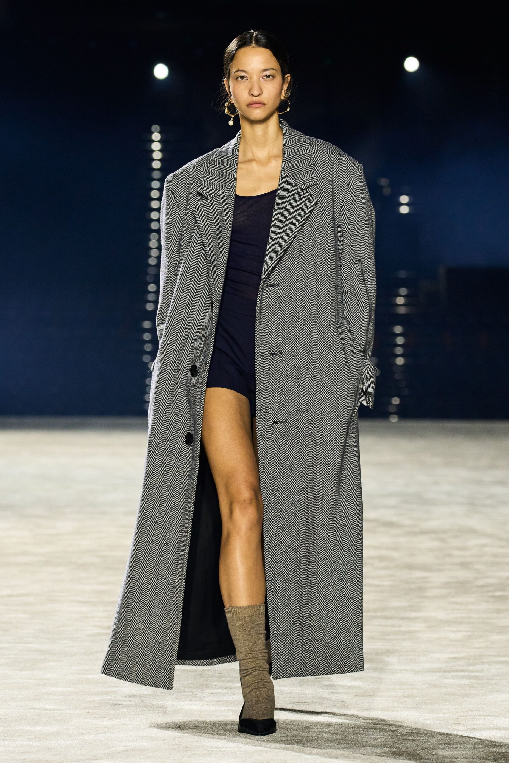 Mismatched gold hoops with a single pearl drop swinging low. Oversized grey herringbone overcoat, broad lapels and one low black button. Inky jersey bodysuit cut to a high bare thigh. Marl taupe socks slouched above the shoe. Black pumps with squared toes. Crossing a hotel lobby at dawn before anyone has checked out.