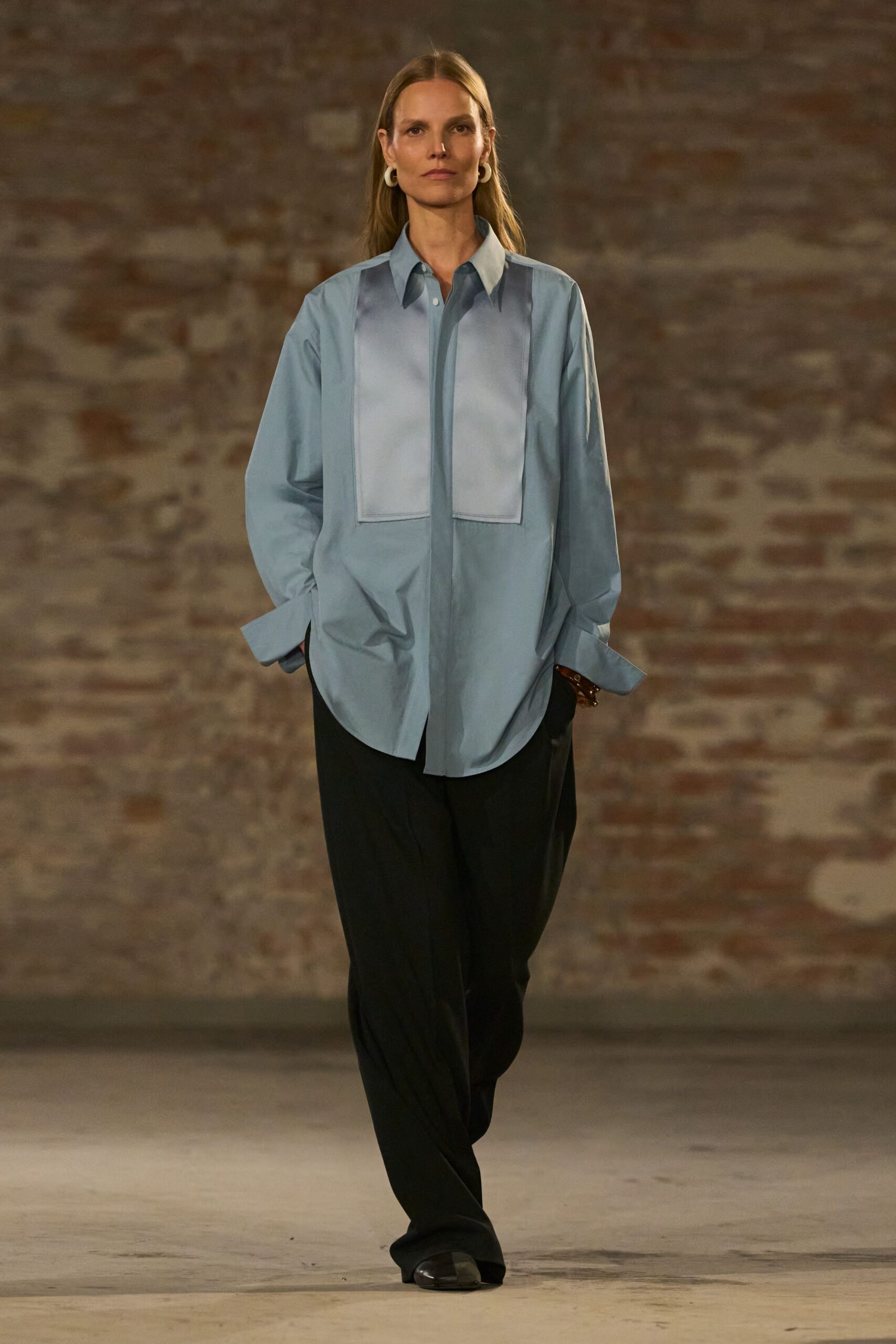 Chunky white hoops hugging the lobes. Powder-blue poplin shirt with a bib-like tonal panel and long cuffs. Black fluid trousers breaking softly over the shoe. Dark square-toe flats with a dull shine. Early meeting in a converted warehouse before the heaters catch up.