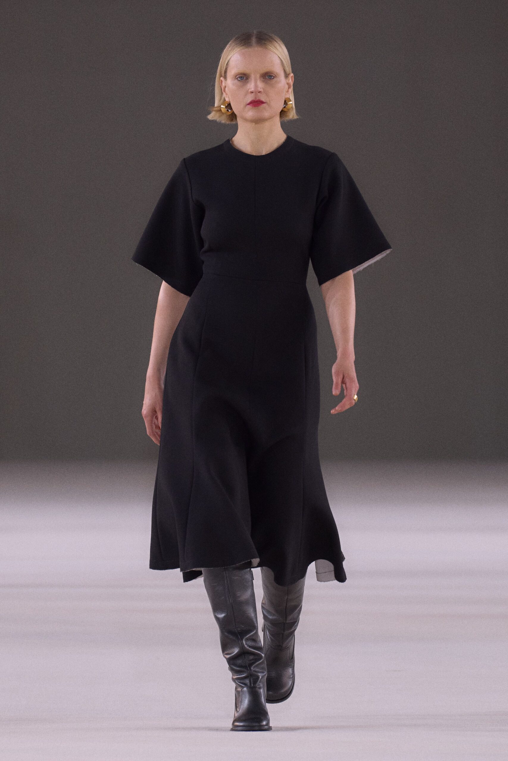 Chunky gold hoops hugging the lobes. Black crepe midi dress with wide elbow sleeves and a center seam. Black leather knee boots, softly creased through the shin. Small gold ring on the right hand. For reading names from a lectern in a hall gone completely still.