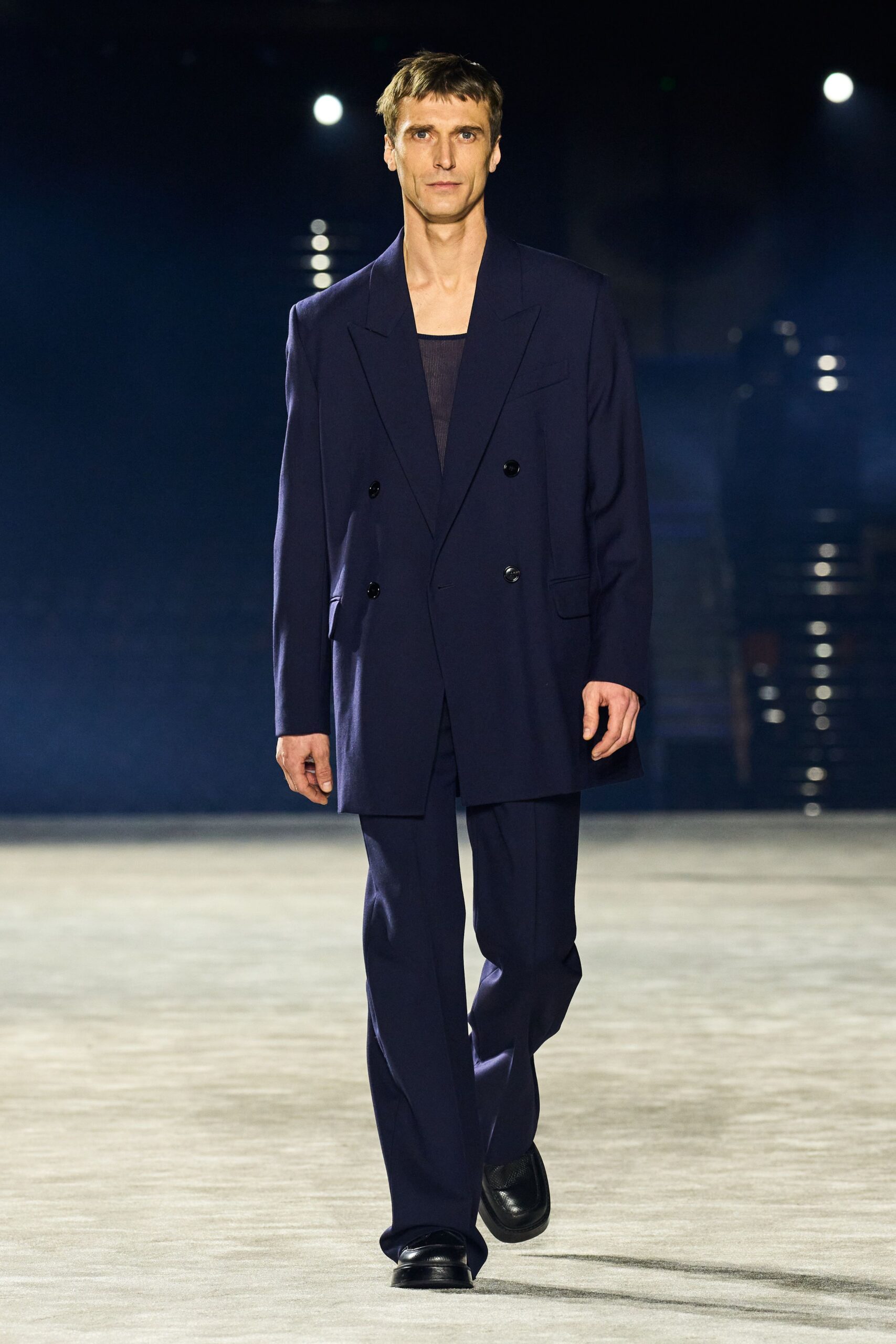 Ink-navy double-breasted blazer cut long, glossy buttons set low. Sheer ribbed tank, square neckline stark under the jacket. Matching straight trousers with a soft break over the shoe. Black leather loafers with squared apron toes. Walking the hotel corridor after midnight ice has melted in the bucket.