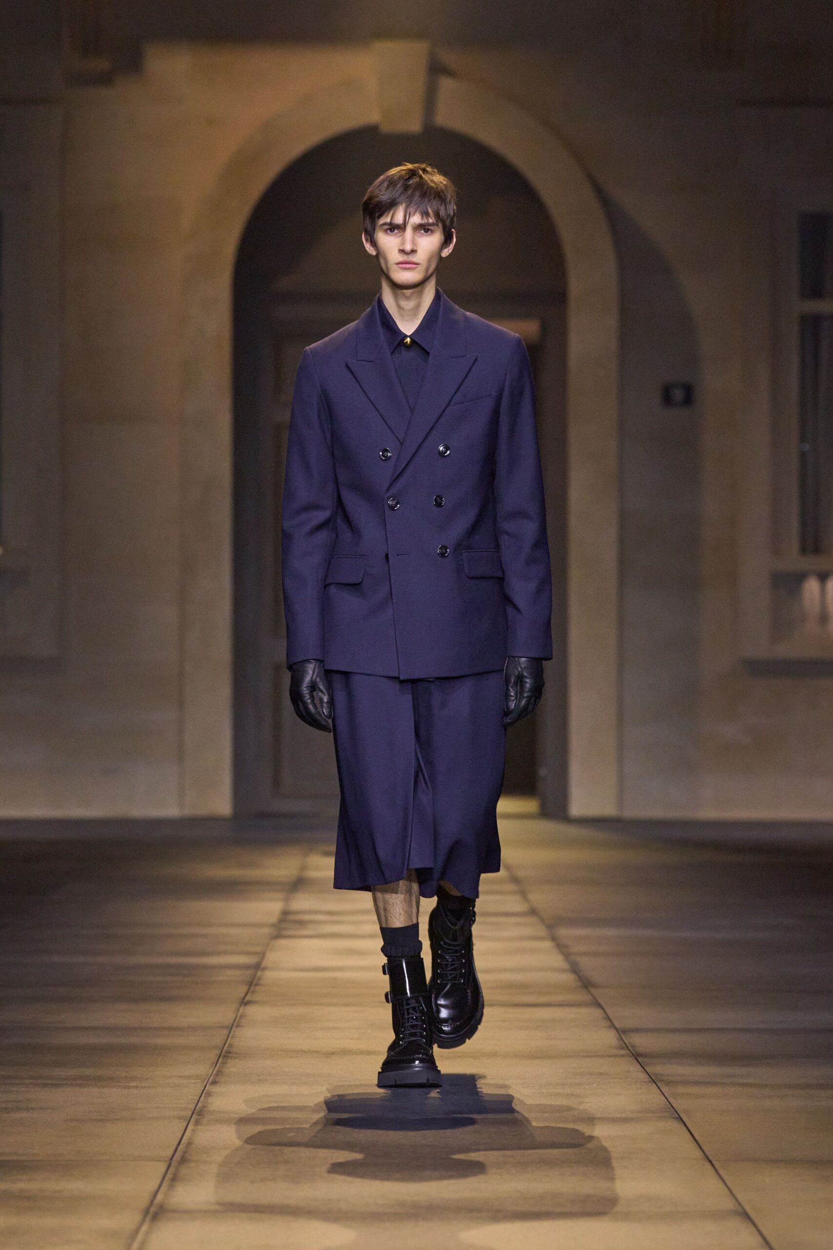 Ink-navy double-breasted blazer with a low six-button stance. Matching shirt fastened to the throat with one small gold stud. Wide cropped trousers breaking below the knee like a divided skirt. Black leather gloves. Glossy combat boots with twin ankle straps. Crossing a stone cloister before the bells finish.