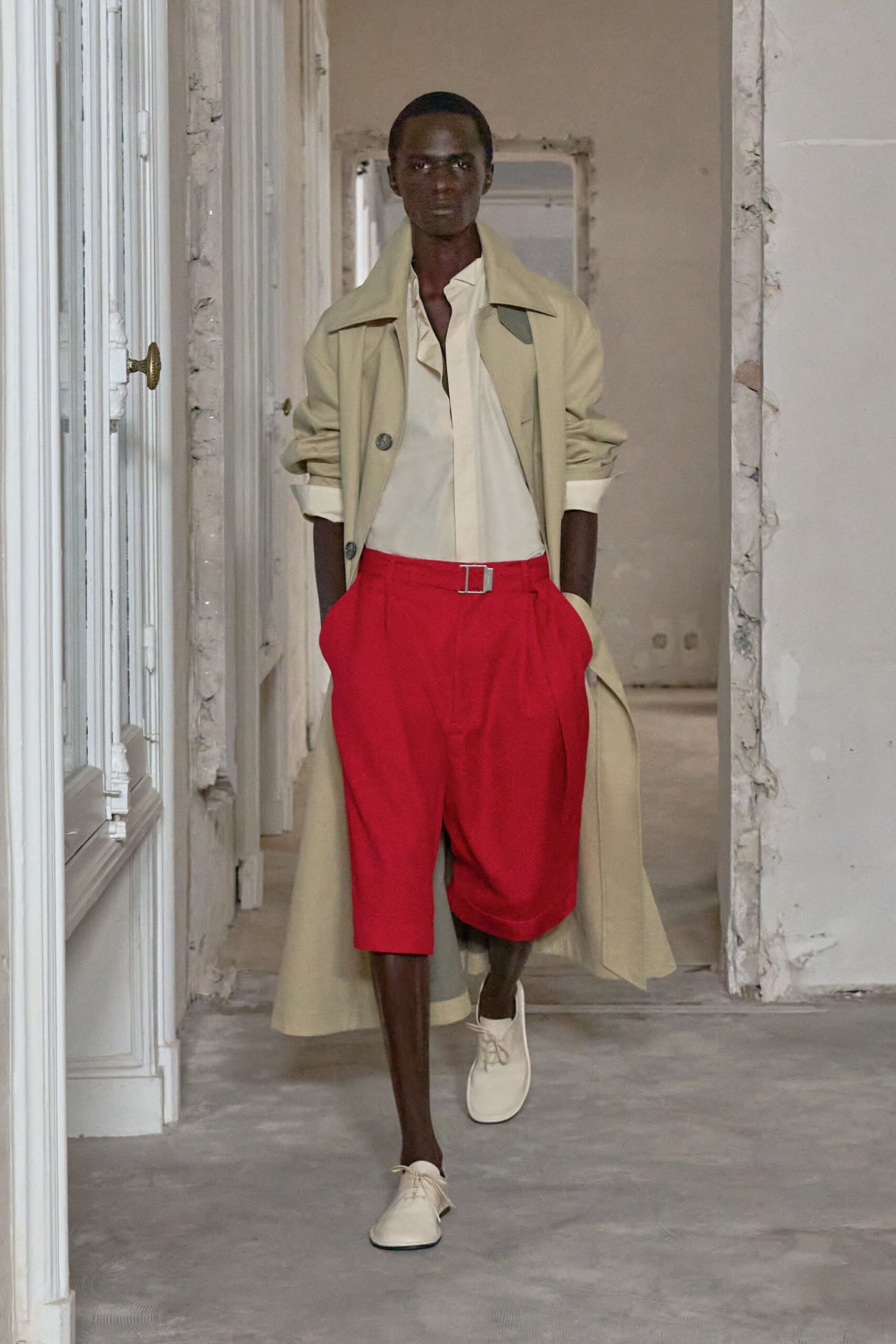 Sand trench coat, sleeves shoved up above clean turned-back cuffs. Cream shirt with a soft placket folding open at the throat. Tomato red pleated shorts, cinched with a square metal-tab belt. Milk-white derbies with plain round laces. Walking through an apartment mid-renovation before the dust sheets arrive.