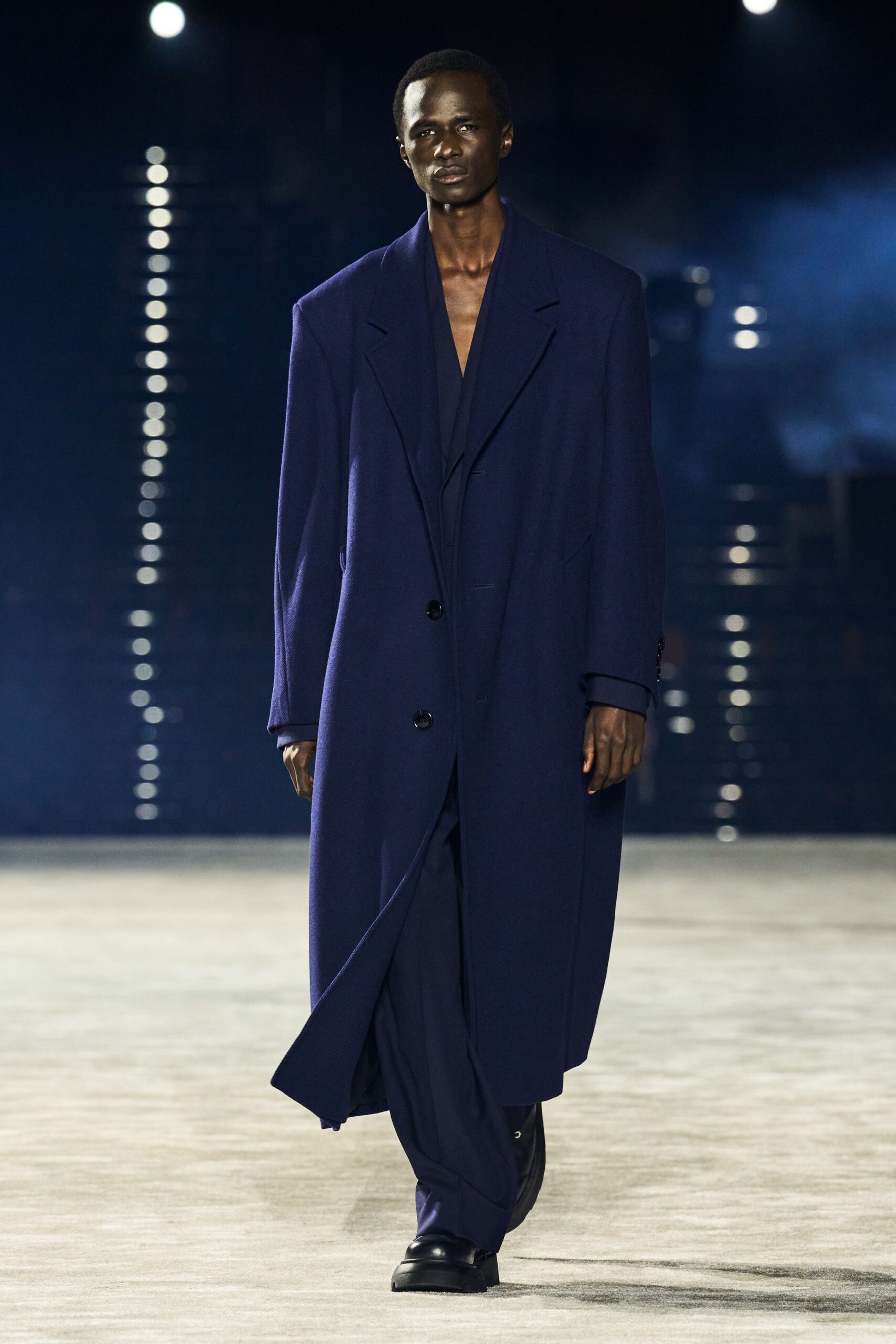 Ink-blue wool overcoat with padded shoulders and low black buttons. Deep V layer beneath, cut almost to the sternum. Matching wide trousers breaking softly over the shoe. Black leather platform derbies. Crossing an empty ferry terminal before the first departure.