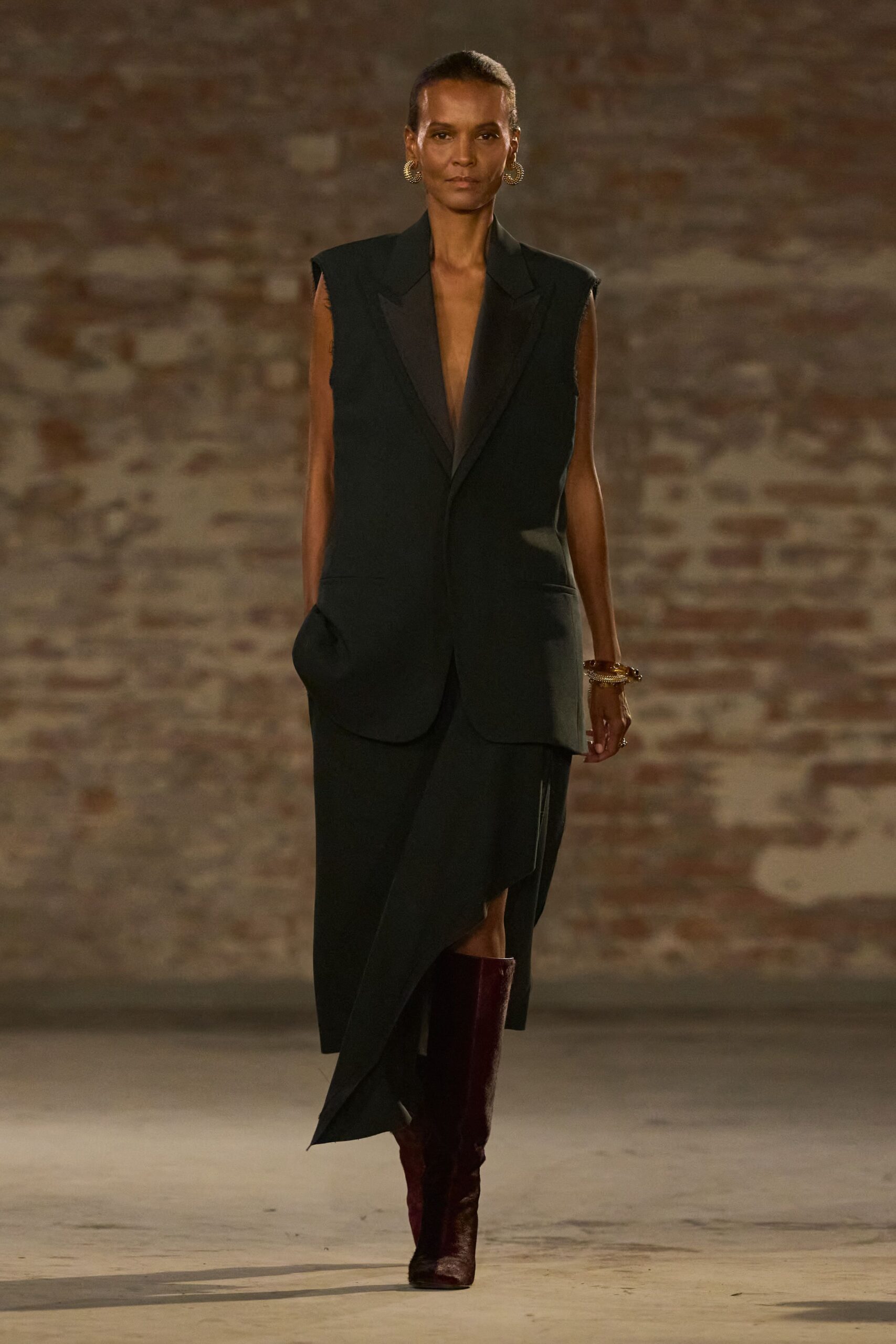 Long black tuxedo vest with satin lapels and raw-cut armholes. Black wrap skirt falling in a sharp apron point across the shin. Dark oxblood knee boots with a glazed, almost mirror finish. Chunky gold rope hoops. Stacked gold bangles at one wrist. For crossing a stone courtyard after midnight with your jacket left behind.