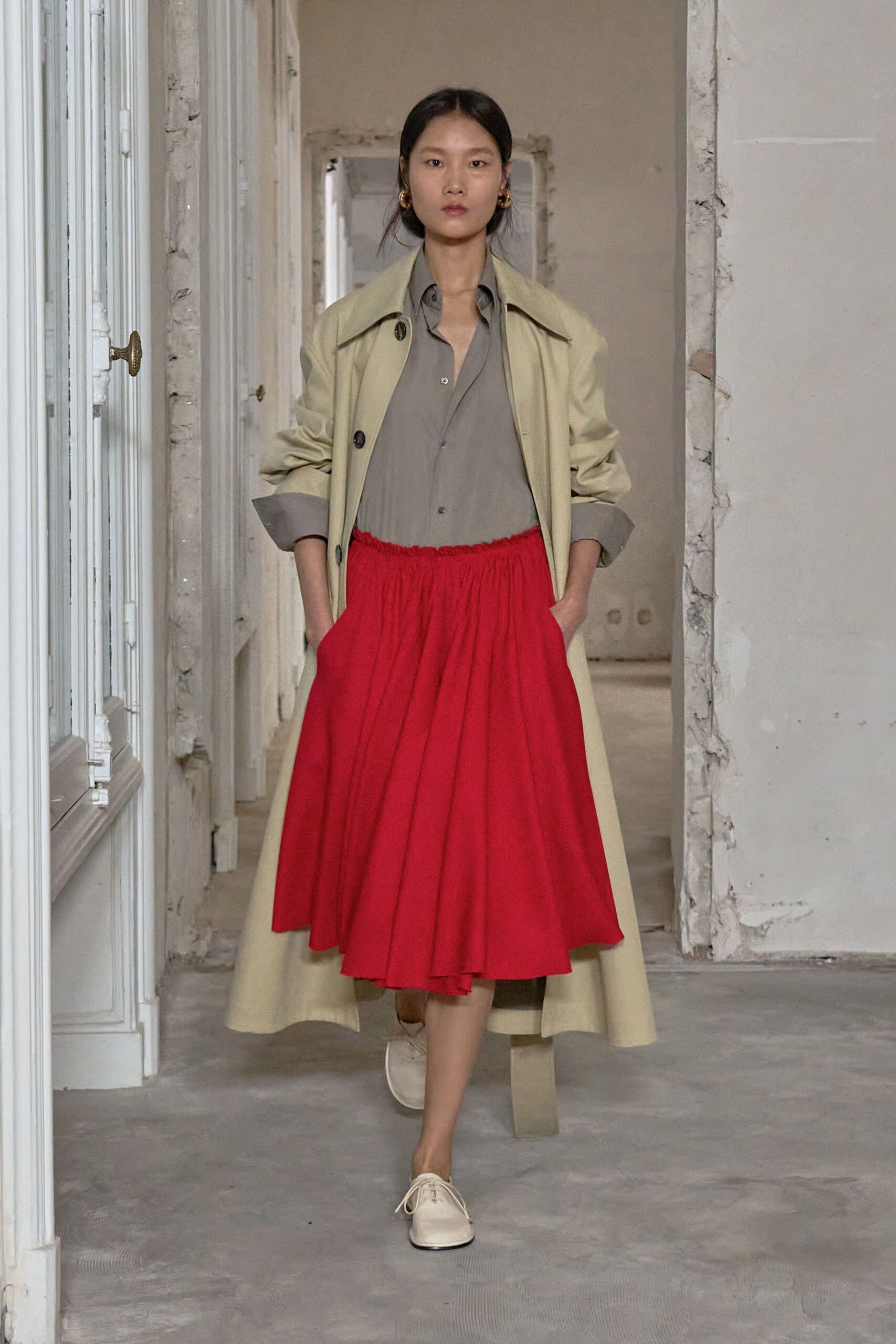 Buttercream coat with broad lapels and sleeves shoved into thick cuffs. Taupe fluid shirt, top buttons left open above tiny smoky buttons. Poppy red gathered skirt, crinkling from a high elastic waist. Cream lace-up oxfords. Crossing a half-renovated hotel corridor before the wedding guests wake.