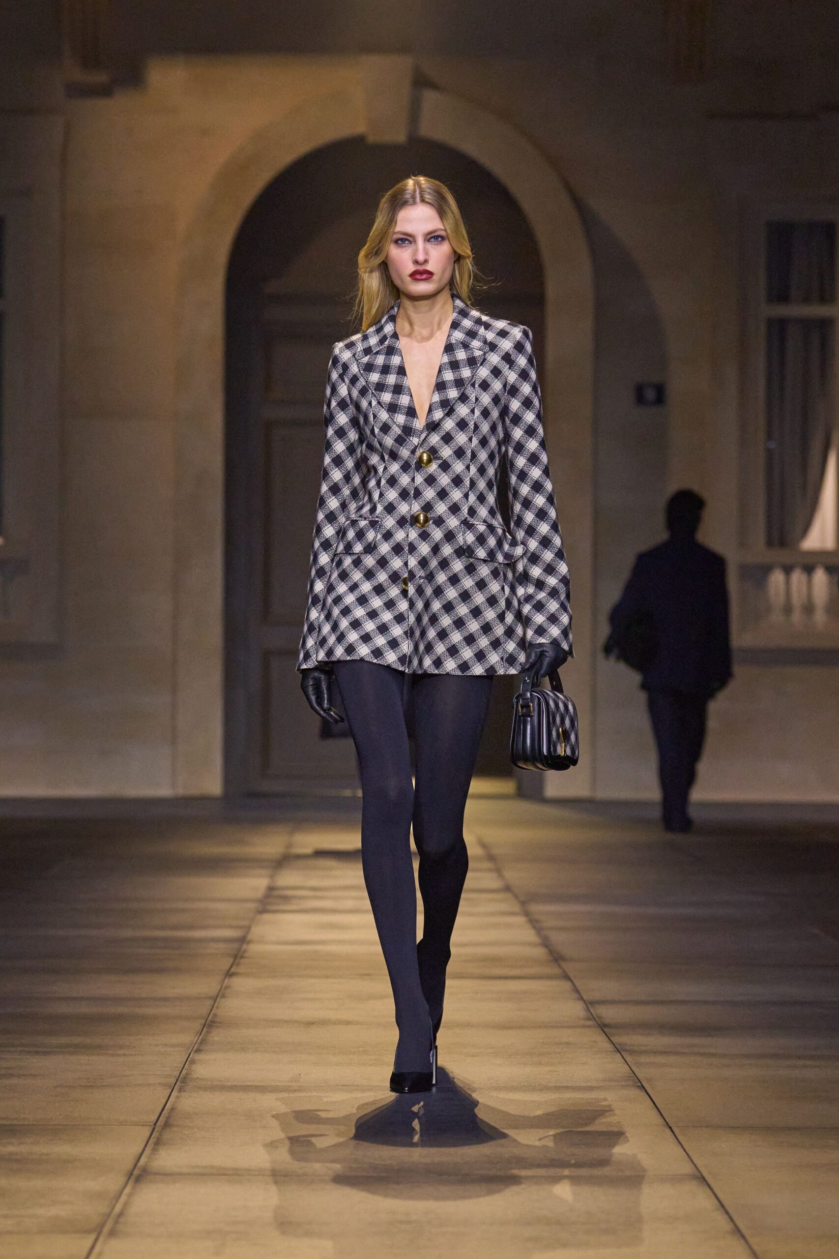 Long black-and-white gingham blazer, cut low with domed brass buttons. Opaque navy tights, unbroken from hem to pointed toe. Black leather gloves. Boxy striped top-handle bag with a gleaming oval clasp. Black patent pumps. Crossing a stone courtyard just before the hotel bar fills for dinner.