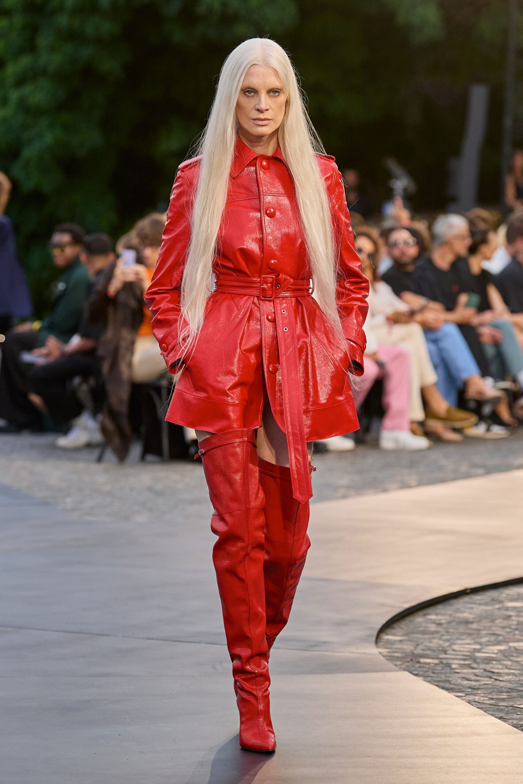 Glossy scarlet leather coatdress with oversized covered buttons and epaulettes. Matching leather belt, long punched tail hanging past the hem. Scarlet thigh-high boots in ribbed leather, buckled at the top. Crossing a hotel courtyard after rain, still carrying the argument with you.