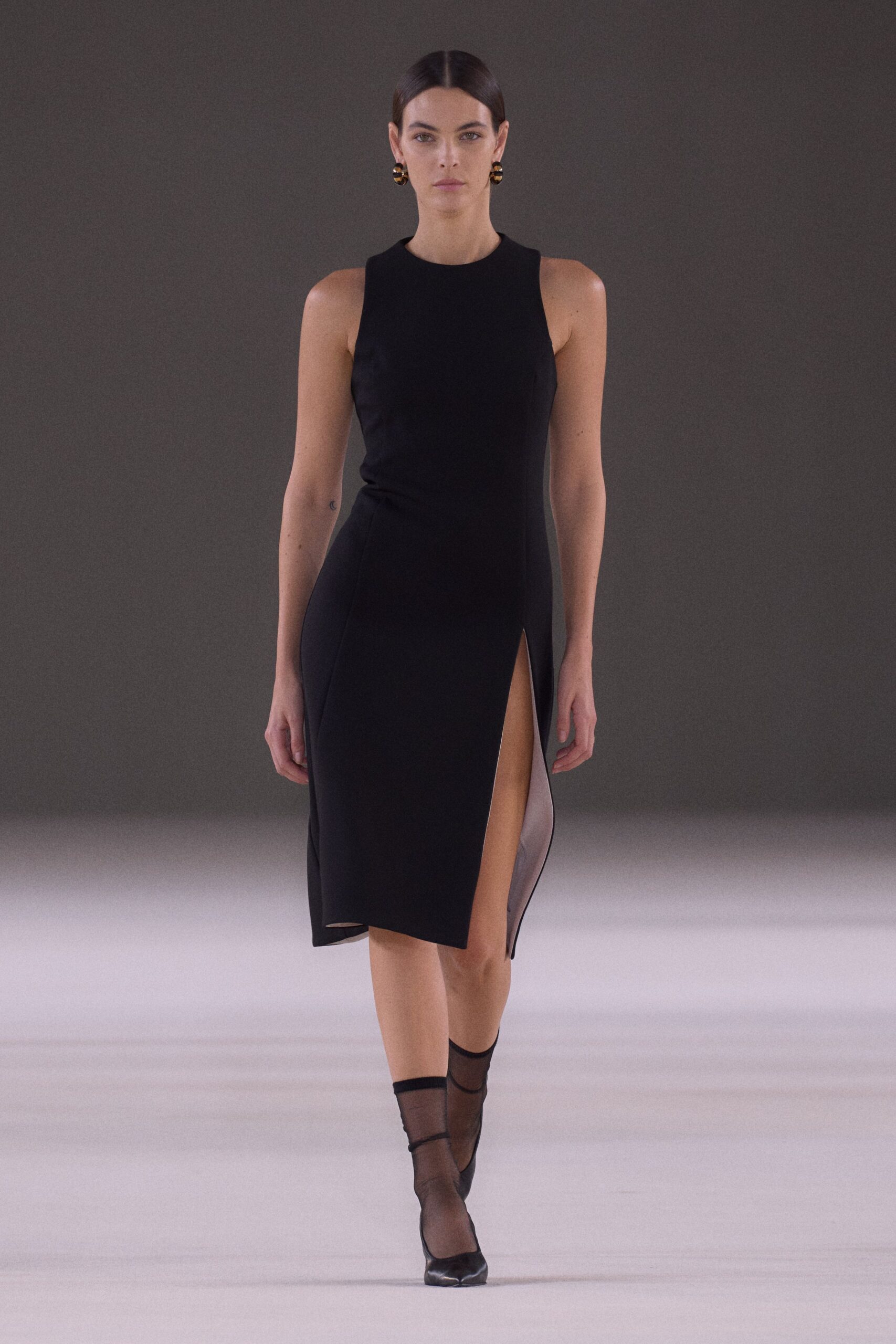 Black crepe sheath dress, cut with a thigh-high slit and sheer underlay. Chunky striped hoops in black and gold. Smoke-black sheer ankle stockings. Black pumps with a split toe seam. For crossing a hotel lobby after midnight with room key in hand.