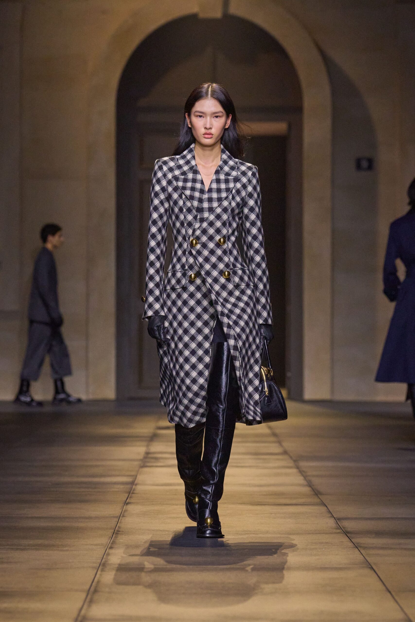 Black-and-ivory gingham coatdress, sharply waisted with brushed brass double-breasted buttons. Black leather gloves, close and unlined. Small black top-handle bag with a curved brass frame. Black leather thigh boots, one brass toe cap catching the light. Crossing a stone courtyard before the hearing starts.