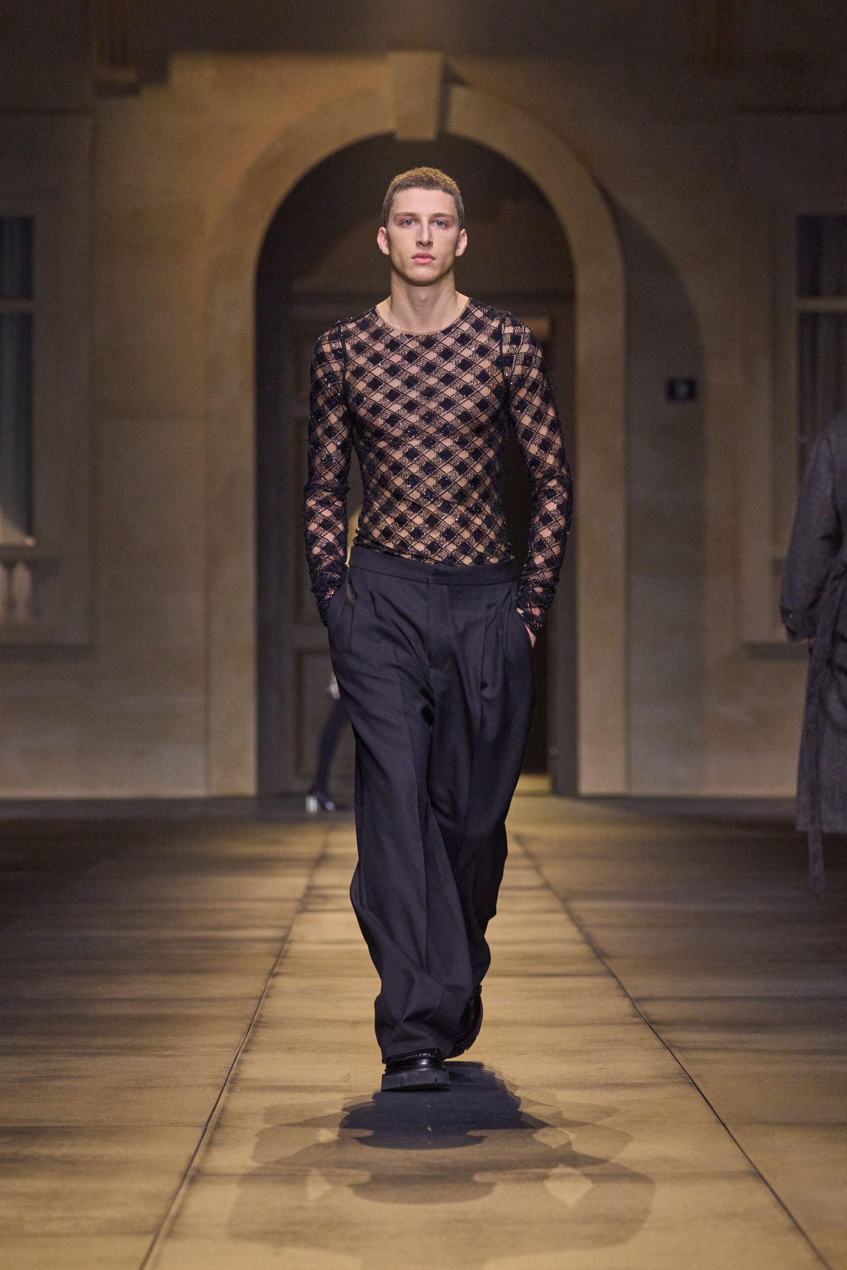 Black openwork mesh top in a diamond lattice over bare skin. Inky pleated trousers cut wide, waistband sitting clean and high. Square-toed black leather shoes with a low blunt shine. Crossing a hotel courtyard after midnight, windows still lit upstairs.