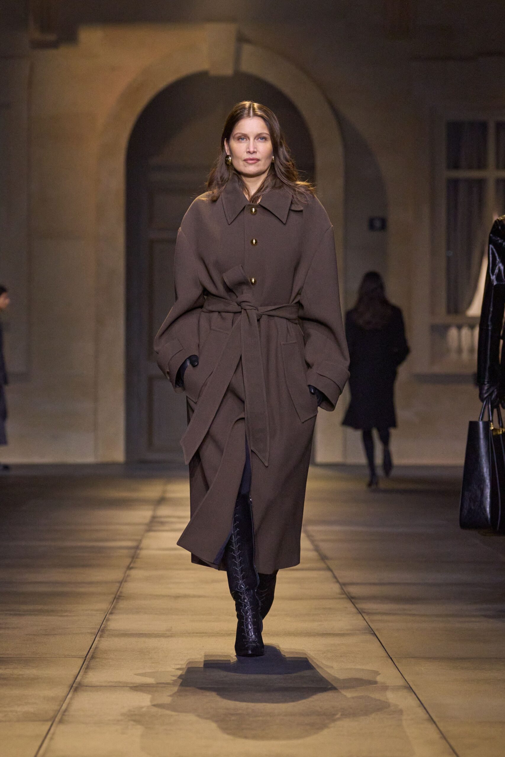 Chocolate wool coat with dropped shoulders, horn buttons and a long self-tie belt. Black knee boots, front-laced tight to the shin. Small round gold hoops. For crossing a stone courtyard before dinner, when the air has turned cold.
