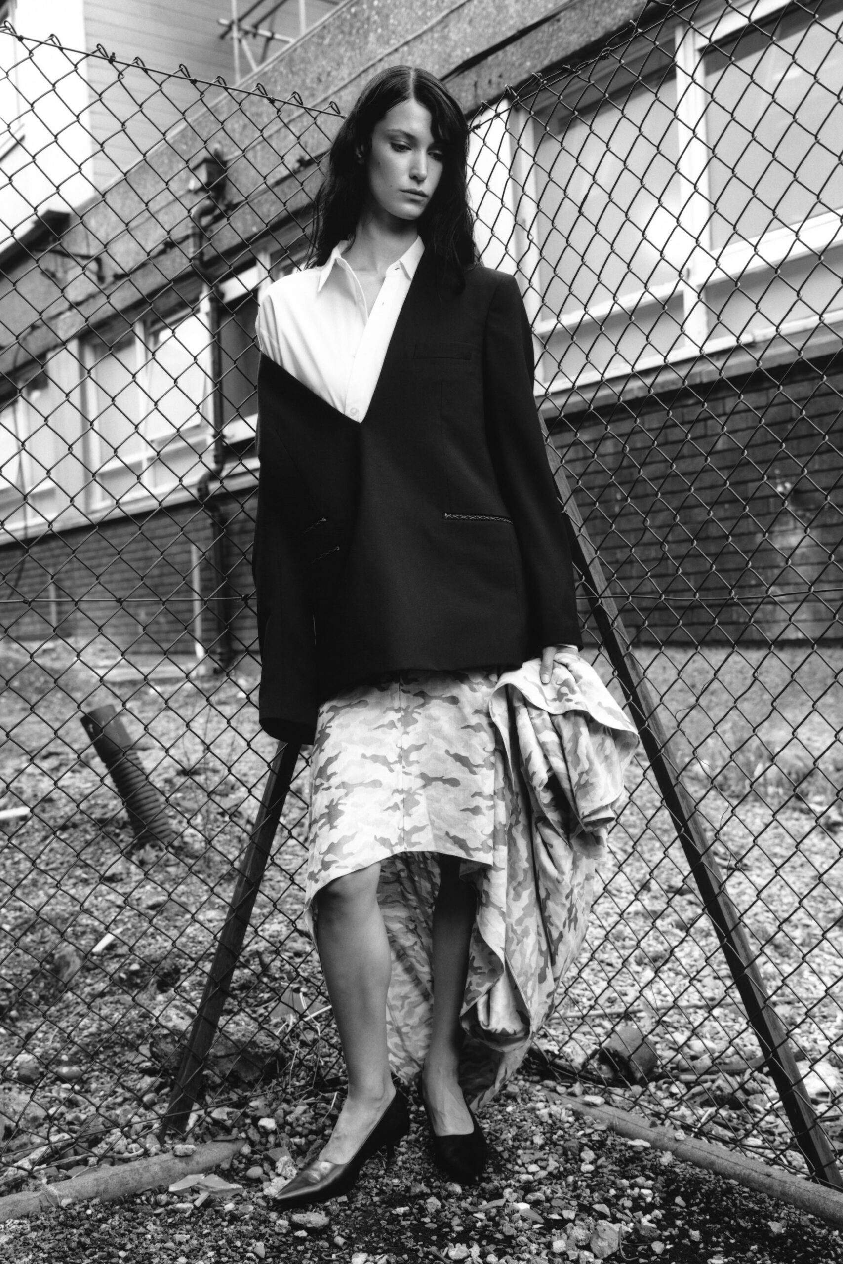 White button-front shirt with a narrow placket and pointed collar. Black blazer cut with one shoulder dropped low off the body. Asymmetric camouflage skirt, one panel trailing crumpled in her hand. Black pointed pumps with a sharp, low-cut vamp. Crossing a fenced-off lot after rain, office clothes gone slightly wrong.