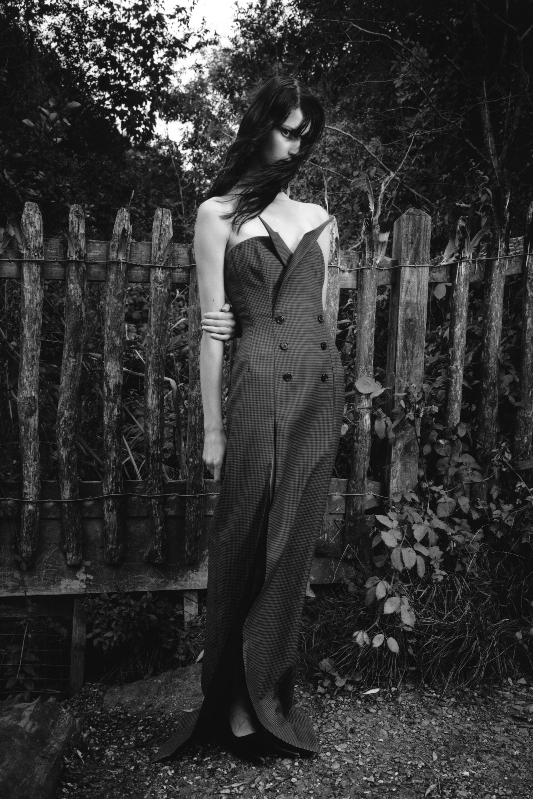 Strapless micro-check jumpsuit cut like a double-breasted coat, peaked lapels folding inward. Black pointed shoes barely visible under the pooling hem. By the split-rail fence after rain, when the path has turned to grit.