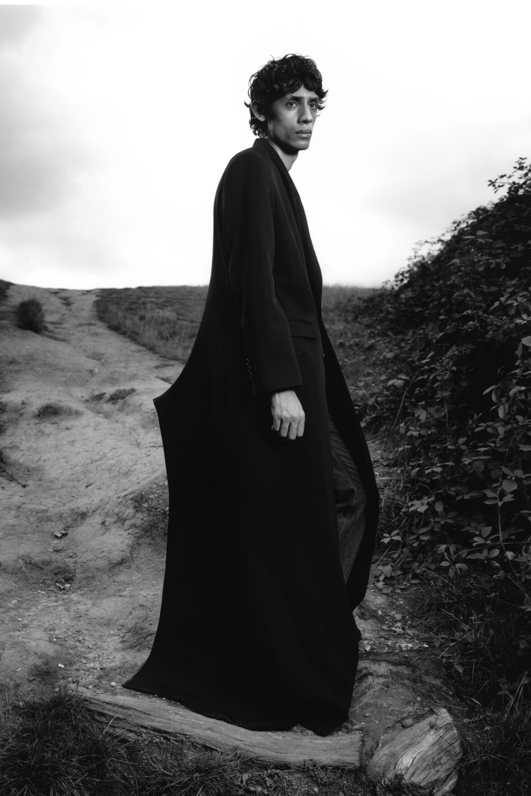 Floor-length black coat in dense wool, open front falling almost to the ground. Dark trousers with a faint sheen, loosening into the coat's sweep. Crossing a scrubby hillside after rain, cuffs skimming the dirt track.