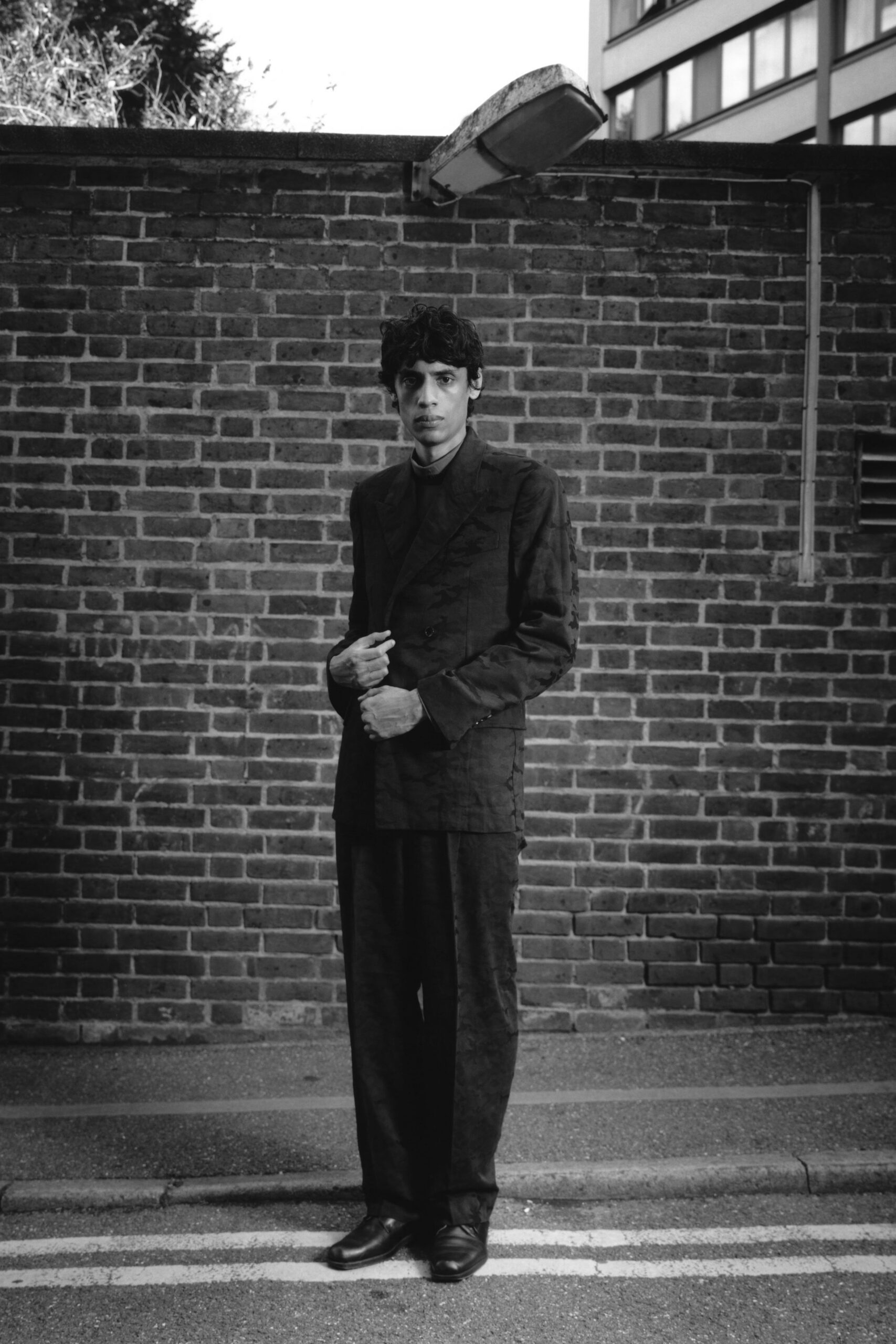 Dark double-breasted jacket in faintly lustrous jacquard, peaked lapels sitting narrow. High-neck underlayer close to the throat. Wide straight trousers in the same shadowy patterned cloth. Square-toe leather shoes, softly creased over the vamp. Crossing a side street after rain, office windows already gone dark.