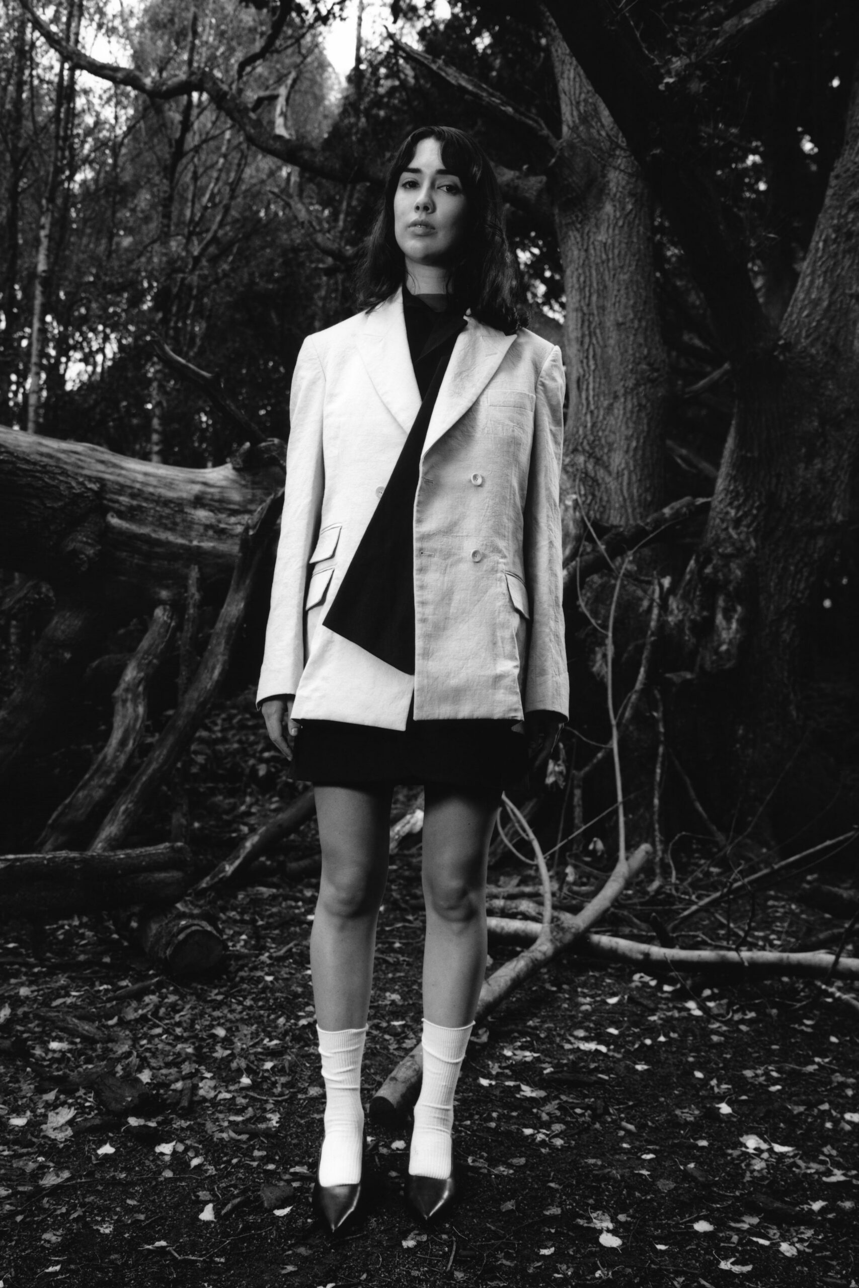Pale double-breasted blazer in slubby linen, one flap pocket split into two. Black draped top falling in a long scarf panel. Short black skirt, hem just clearing the blazer. White ribbed crew socks. Dark pointed pumps with a sharp vamp. Walking through wet woodland after changing out of office clothes.