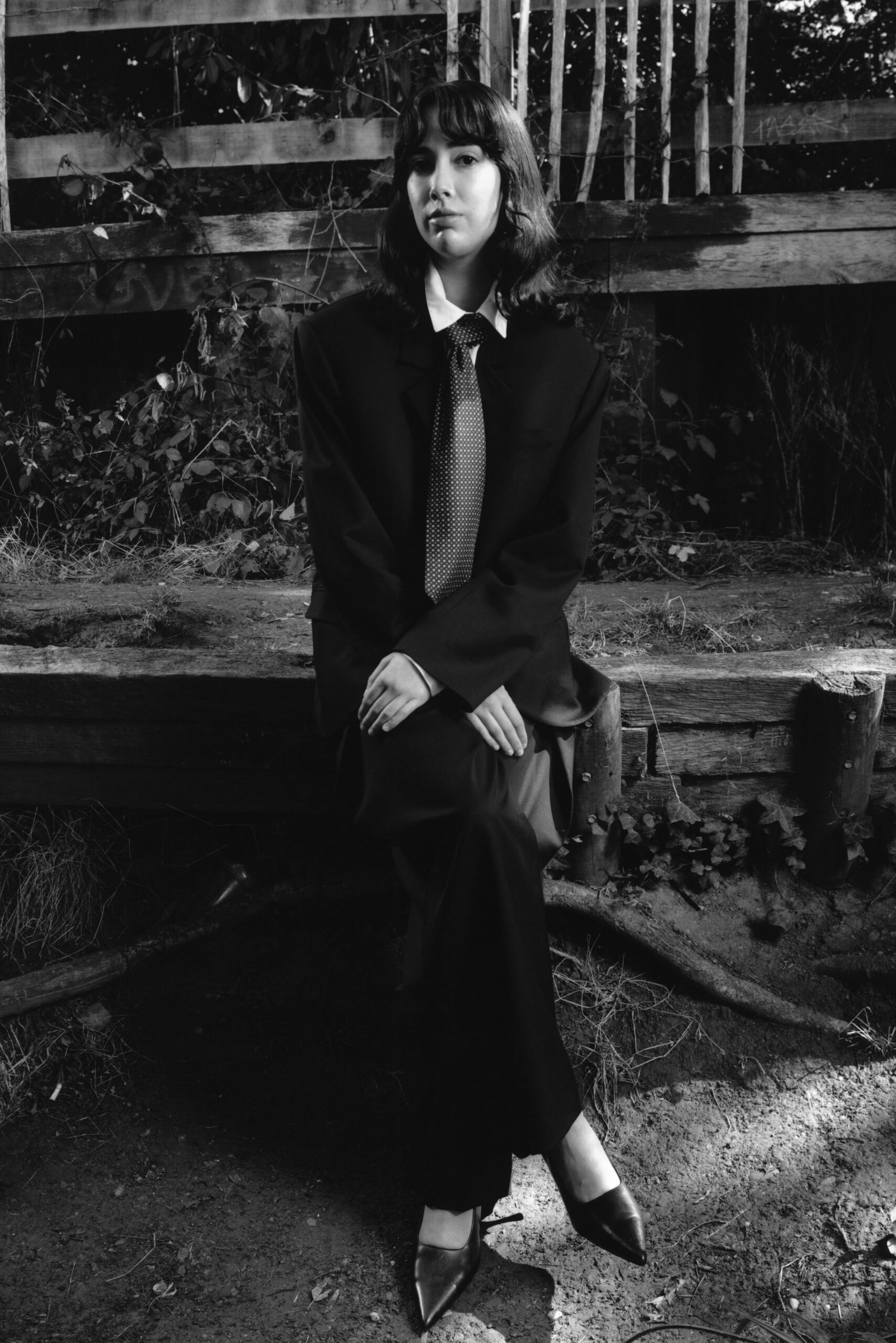 Black tailored blazer with broad shoulders and a low two-button stance. White shirt collar cut wide and crisp over the lapels. Small-dot necktie hanging straight to the waist. Black trousers falling long with a soft break at the shoe. Pointed leather pumps. Sitting by a splintered garden stage after the school play emptied out.