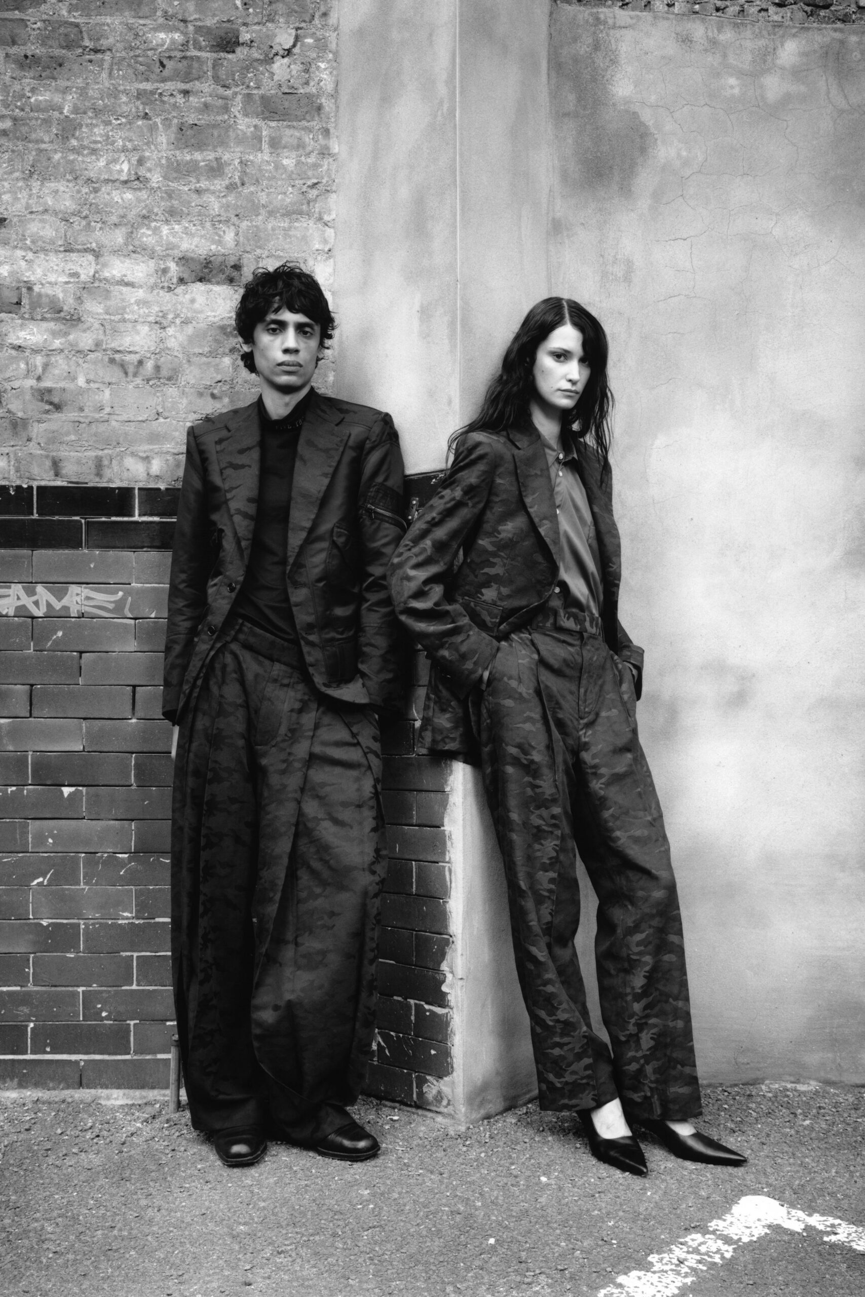 Dark moire blazer with peaked lapels and a nipped waist. Soft plain blouse, top button left casually open. Matching moire trousers cut long and loose through the leg. Black pointed pumps. Outside the registry office after rain, neither of you speaking first.