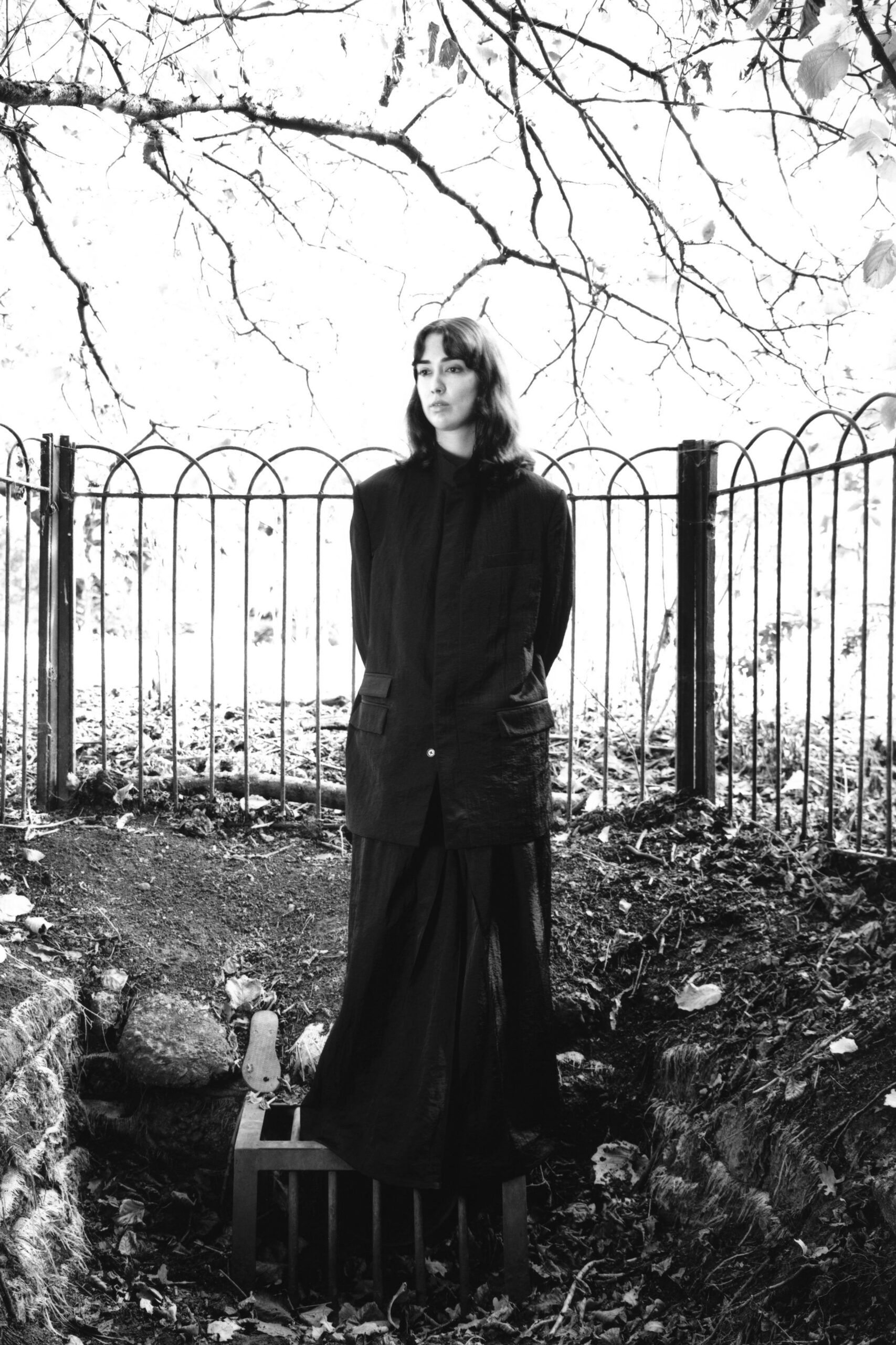 Long black blazer in crinkled cloth, flap pockets set low and square. Floor-length black skirt falling in soft collapsed pleats below the hip. Standing by a fenced creek after rain, leaves plastered to the bank.