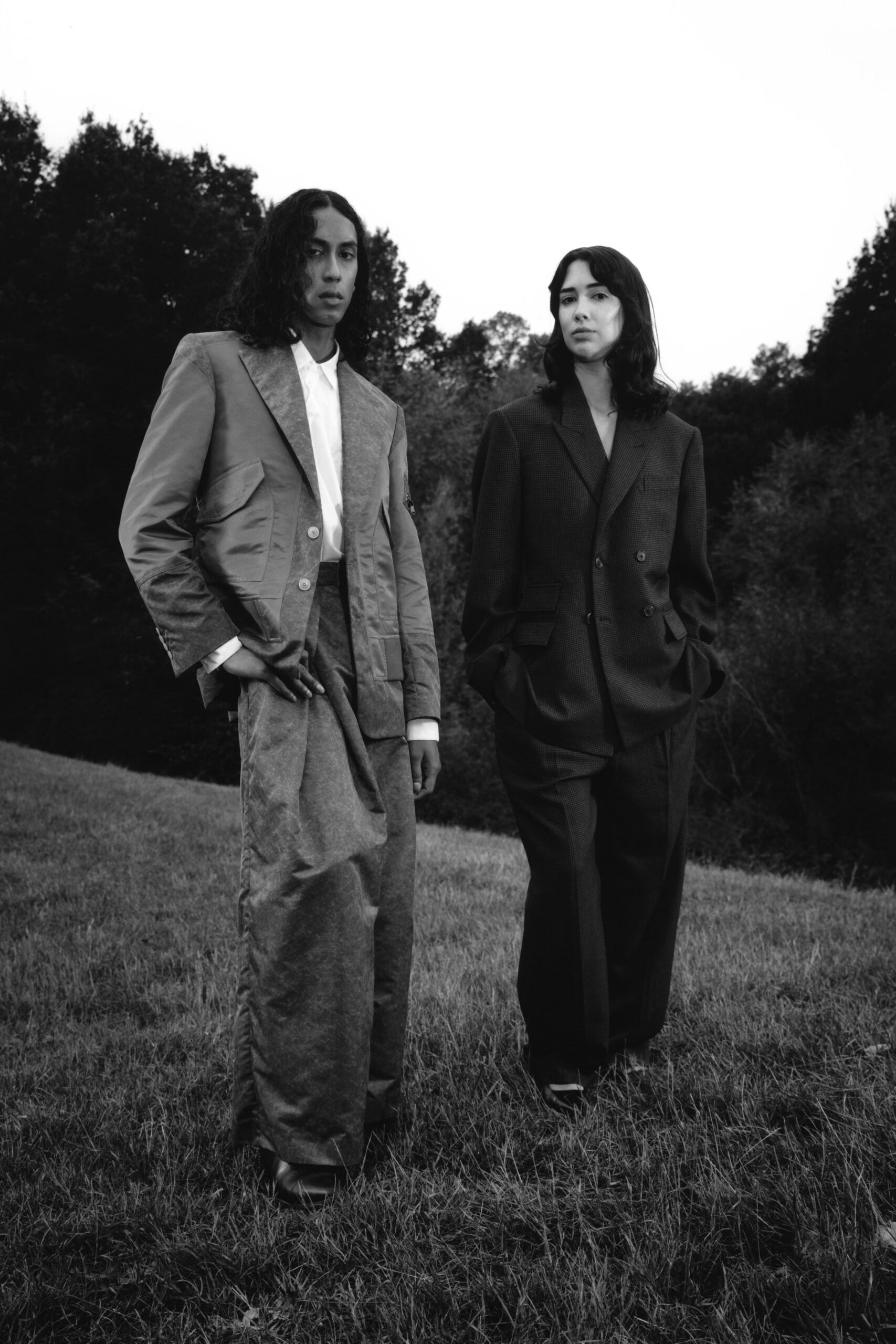 Wide-lapeled blazer spliced from crisp suiting and mottled plush panels. White placket-front shirt, cuffs peeking clean below the sleeves. Loose straight trousers in the same crushed, light-catching cloth. Square-toe leather boots. Standing in wet grass before a country house argument starts.