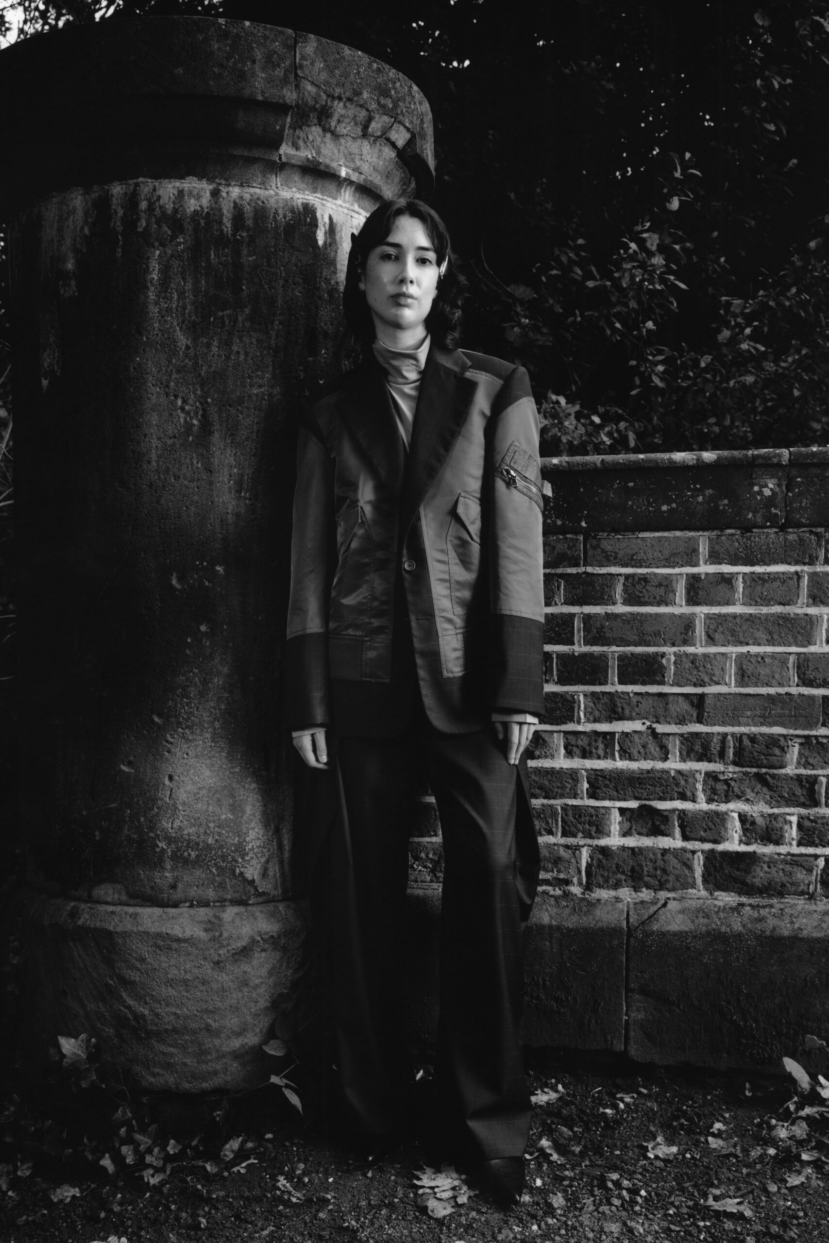 Thin jersey turtleneck gathered softly at the throat. Patchwork blazer with satin lapels, zip pocket strapped to one sleeve. Wide trousers in a faint windowpane check, breaking over the shoes. Pointed dark shoes. By the cemetery wall after rain, before anyone else arrives.