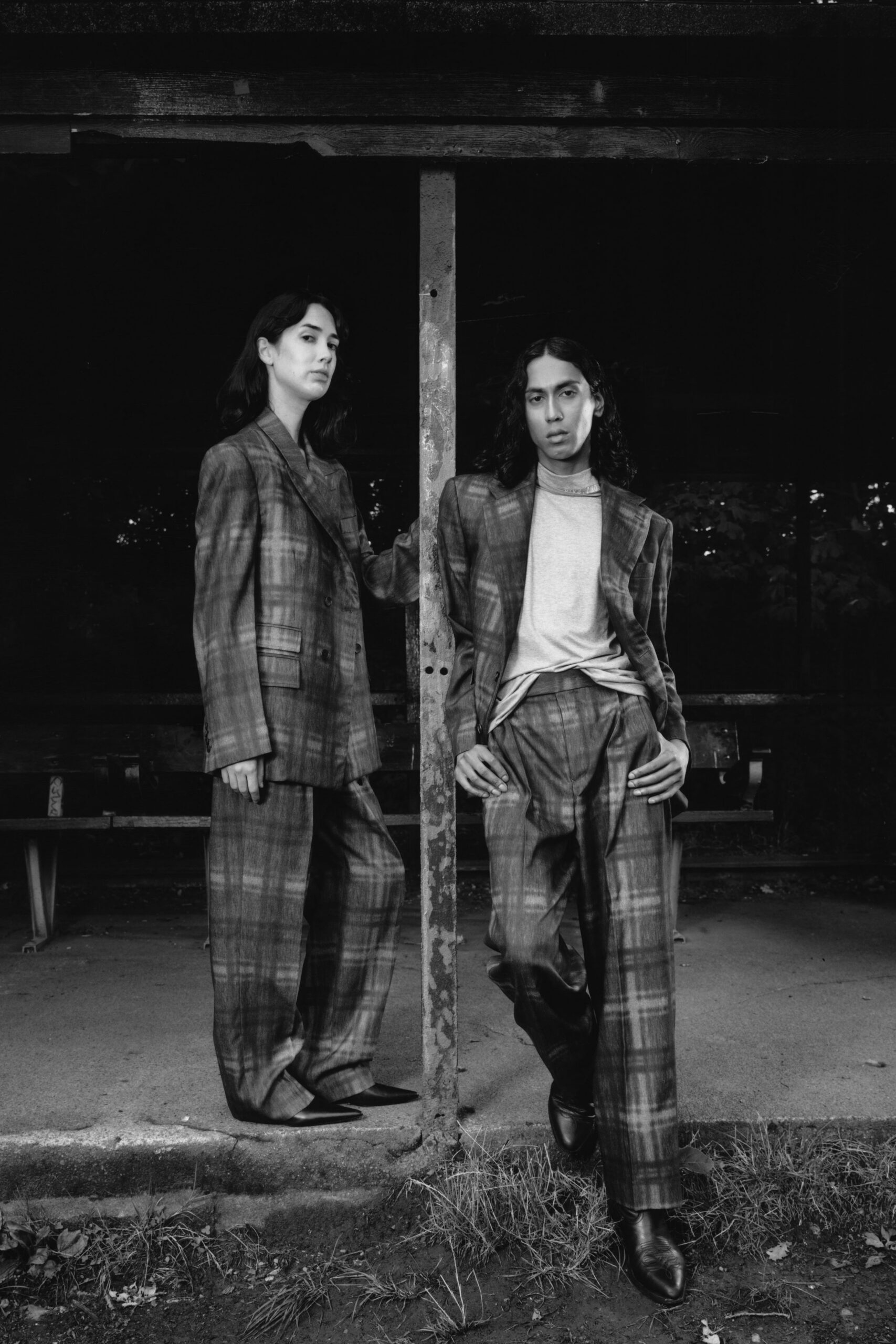 Oversized plaid blazer with broad notch lapels and flap pockets. Pale crewneck tee, hem tugged loose over the waistband. Matching plaid trousers cut wide and softly breaking at the shoe. Black leather ankle boots with squared, narrowed toes. Waiting out drizzle under a park shelter after the last bus left.