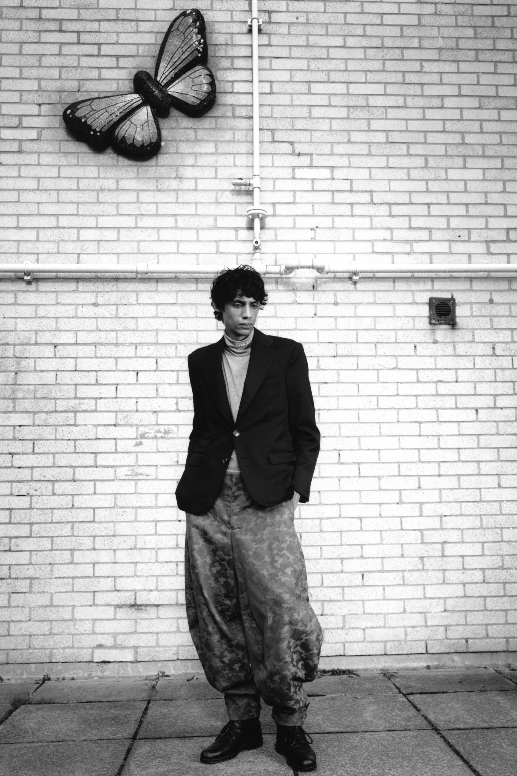 Dark two-button blazer with broad notch lapels and low flap pockets. Slim high-neck knit with tiny lettering circling the folded collar. Balloon trousers in cloudy jacquard, tapering tight at the ankle. Black lace-up boots with plain rounded toes. Outside the museum annex, smoking break beneath a mosaic butterfly.