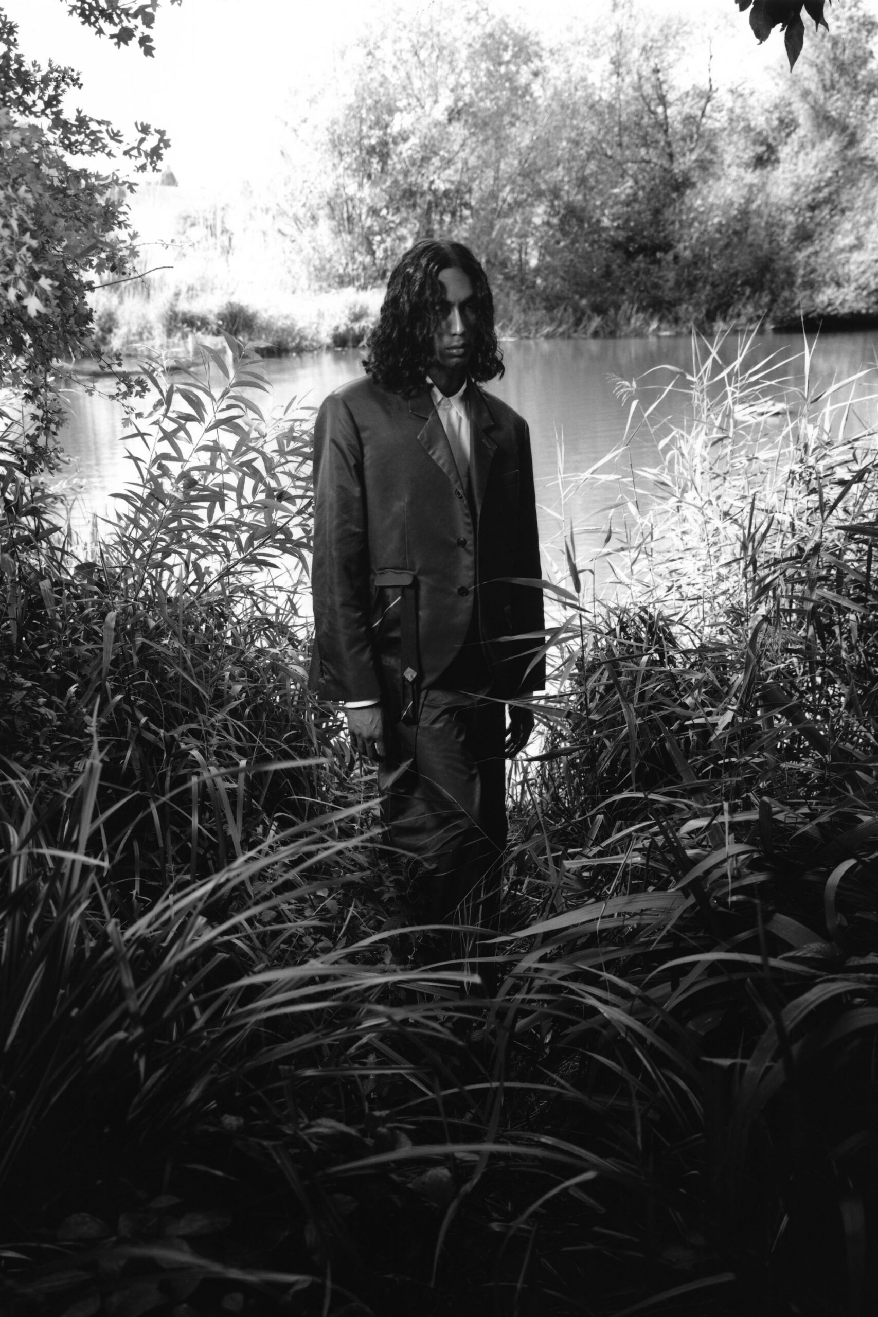 Dark satin-faced blazer with low button stance and flap pockets. Pale shirt buttoned high, narrow collar points tucked under the lapels. Matching tailored trousers with a soft break over the shoe. Standing in reeds at the lake edge after slipping away from the wedding tent.