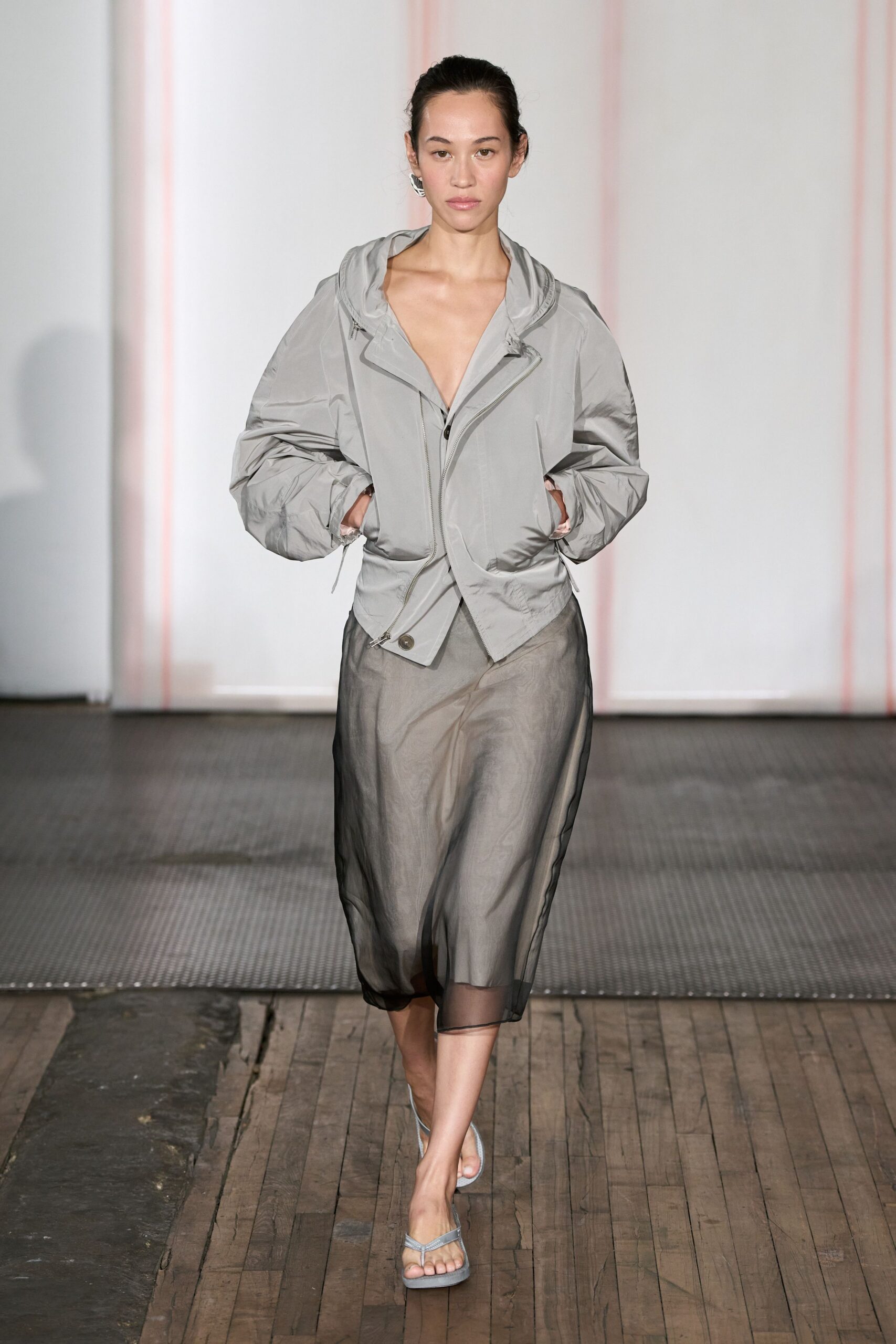 Pale grey nylon hooded jacket with a skewed zip and one exposed snap. Smoky sheer organza slip skirt, the underlayer fading darker at the sides. Powder grey thong sandals with narrow crossed straps. Walking the harbor path after rain, wind still trapped in the hood.