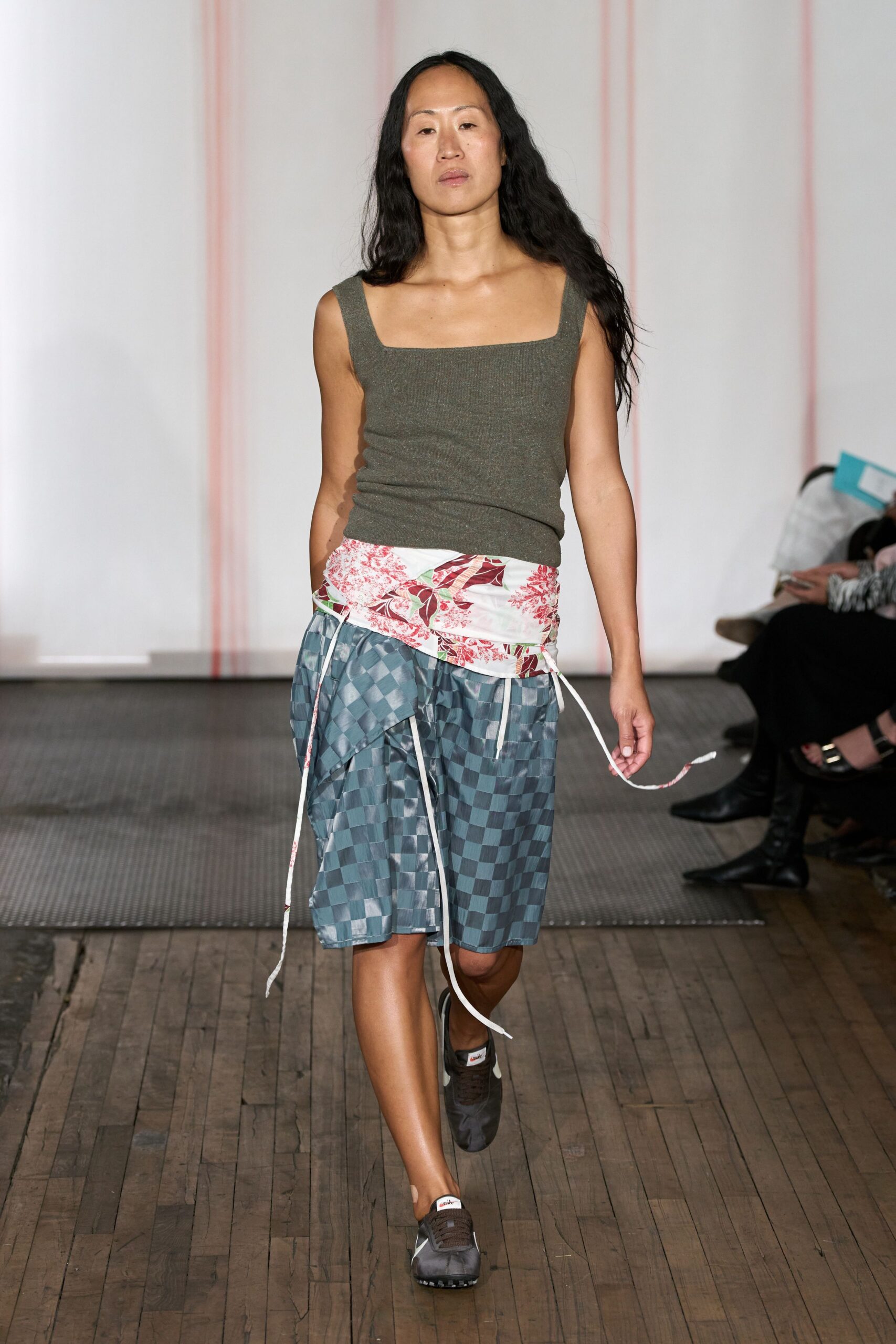 Olive fine-rib knit tank with a squared, ballet-flat neckline. White floral wrap panel tied low, cords trailing past the hip. Blue gray checker satin shorts with a softly puckered surface. Brown nylon runners with white tabs at the tongue. Walking back from the corner store with flowers tucked under one arm.