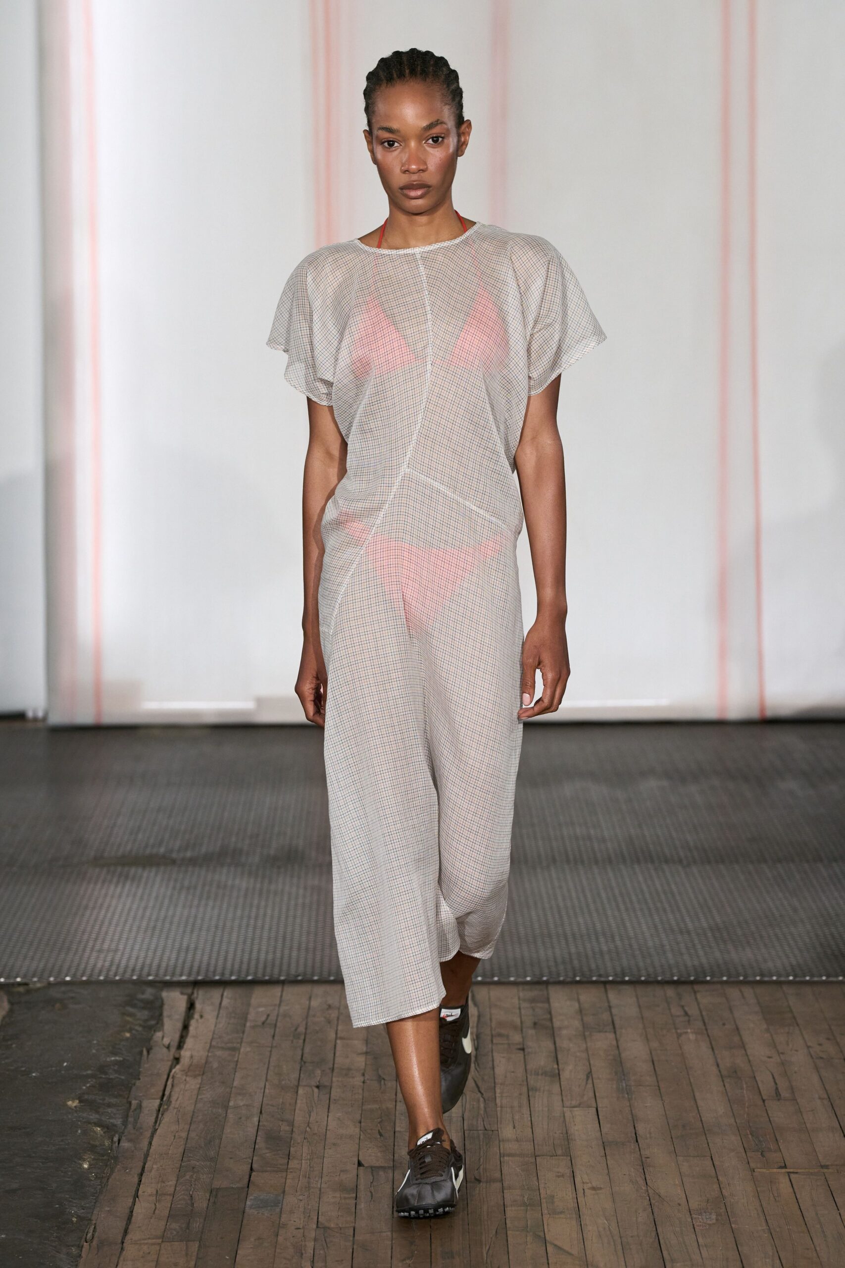 Pale micro-check T-shirt dress, bias seam twisting the body off-center. Neon coral triangle bikini glowing through the semi-sheer weave. Black track sneakers with white side swooshes and waffle soles. Walking from the beach to buy ice before the sun drops.
